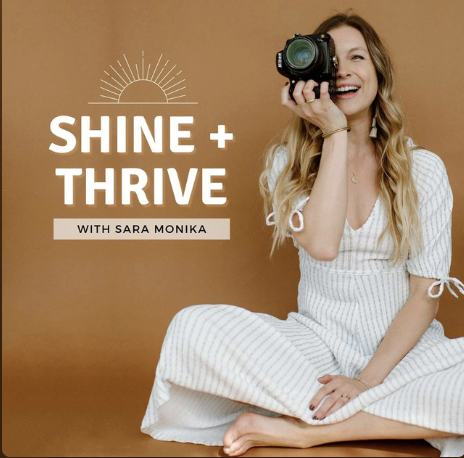A woman with long wavy blonde hair, wearing a white striped dress, sitting cross-legged on the floor, smiling and holding a camera to her eye. The background is a warm brown color with white text that reads 'SHINE + THRIVE WITH SARA MONIKA' and a simplistic sun illustration.