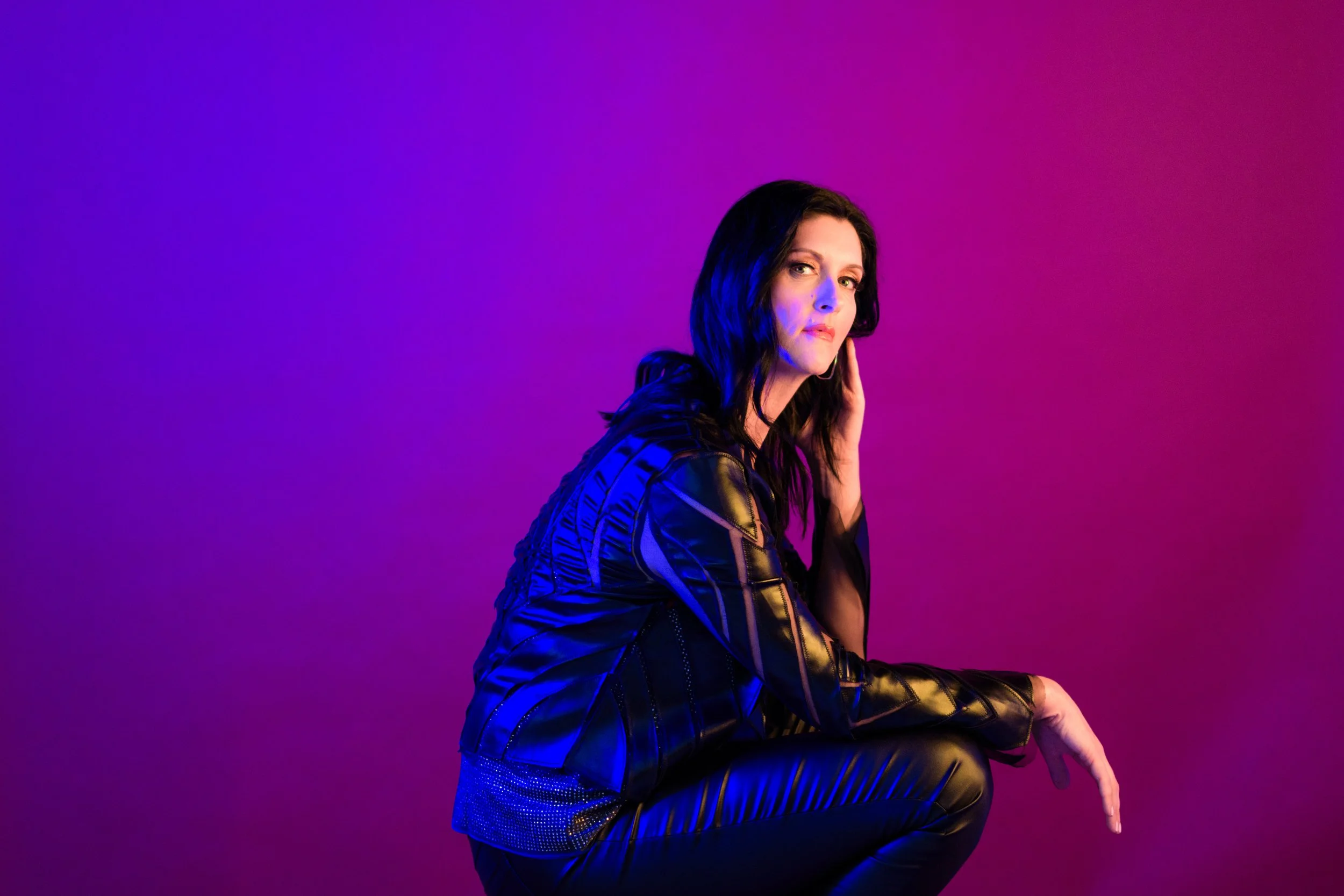 A woman with dark hair, wearing a black leather jacket, squatting against a pink and purple gradient background.