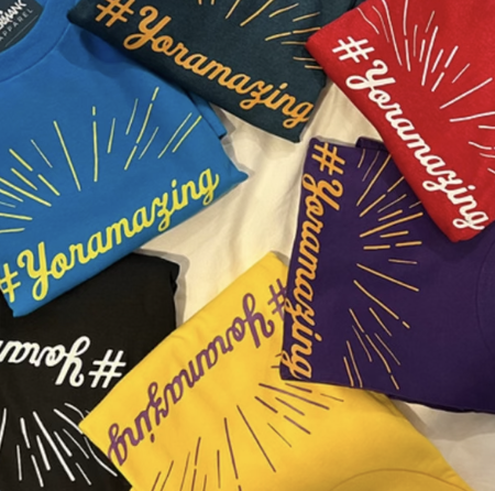 Colorful T-shirts in blue, black, yellow, red, and purple with the hashtag #Yonamazing and graphic lines.