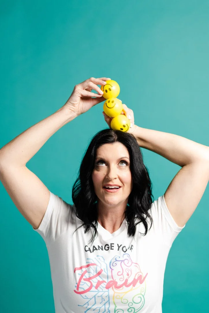 A woman, Jaime Armstrong, with black hair wearing a white t-shirt with colorful text and graphics, balancing three yellow stress balls with smiley faces on her head against a teal background.