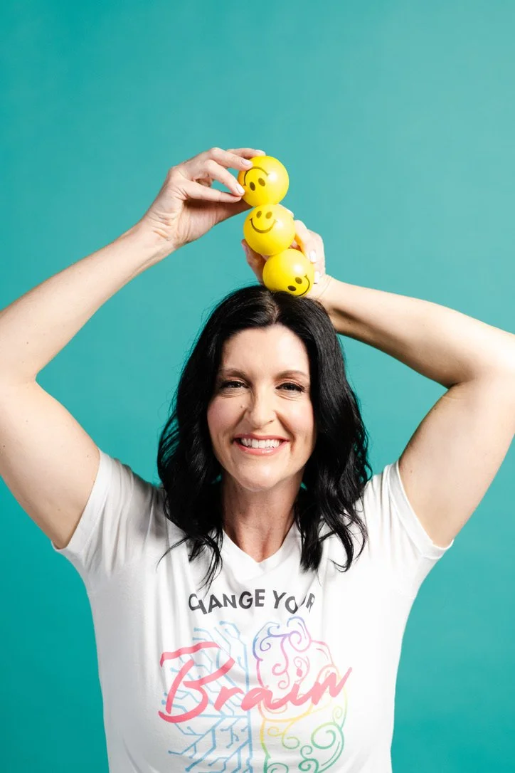 Woman, Jaime Armstrong, with black hair smiling, holding three yellow balls with smiley face emojis above her head against a turquoise background, wearing a white t-shirt with colorful text.
