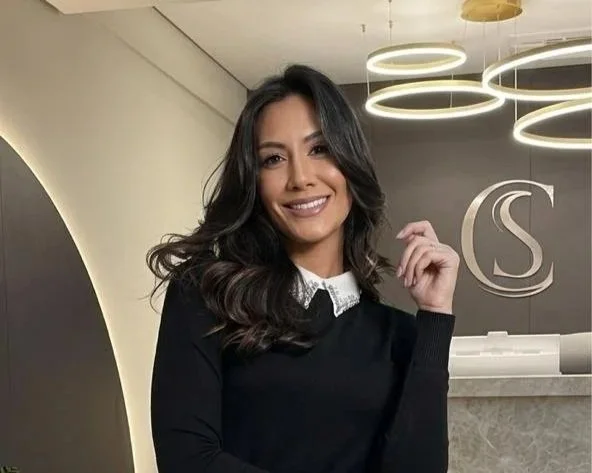 Mulher sorridente com cabelo castanho escuro ondulado, usando suéter preto e camisa branca com gola, em ambiente moderno com decoração de iluminação circular no teto.