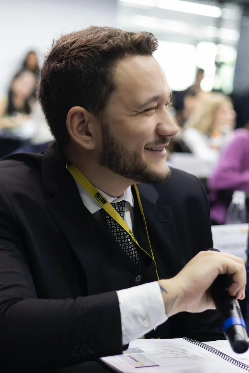 Homem sorrindo em um ambiente de escritório ou conferência, com outras pessoas ao fundo, usando terno, uma fita lanyard amarela e segurando um microfone.