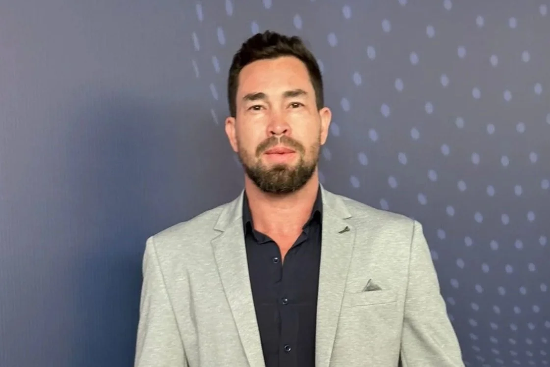 Homem com barba, usando terno cinza e camisa preta, diante de fundo azul com pontos de luz.
