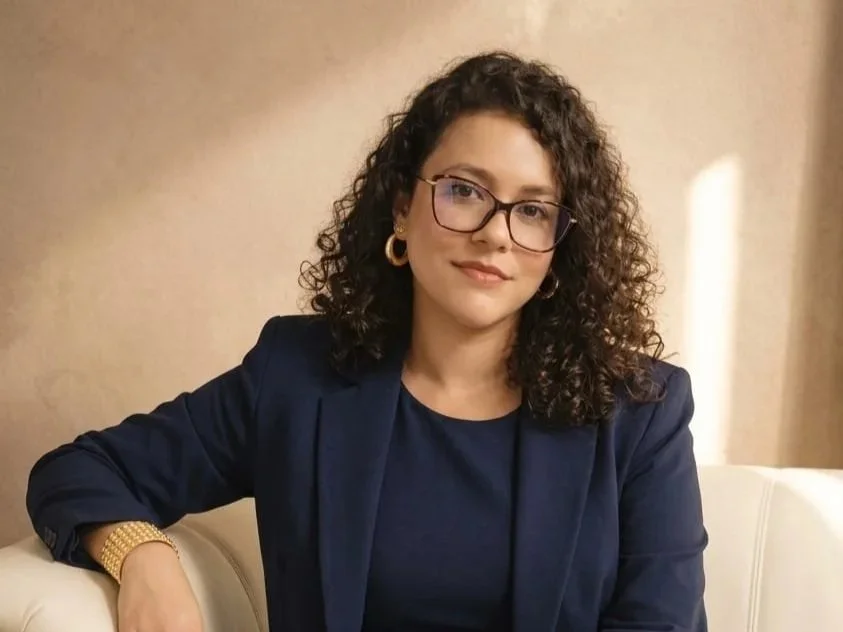 Mulher com cabelo cacheado, usando óculos, brincos dourados, blazer azul e blusa azul, sentada em sofá com fundo de parede de cor clara.