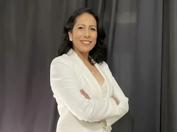 Mulher sorridente com braços cruzados, vestindo blazer branco, em ambiente interno ao lado de cortinas pretas.