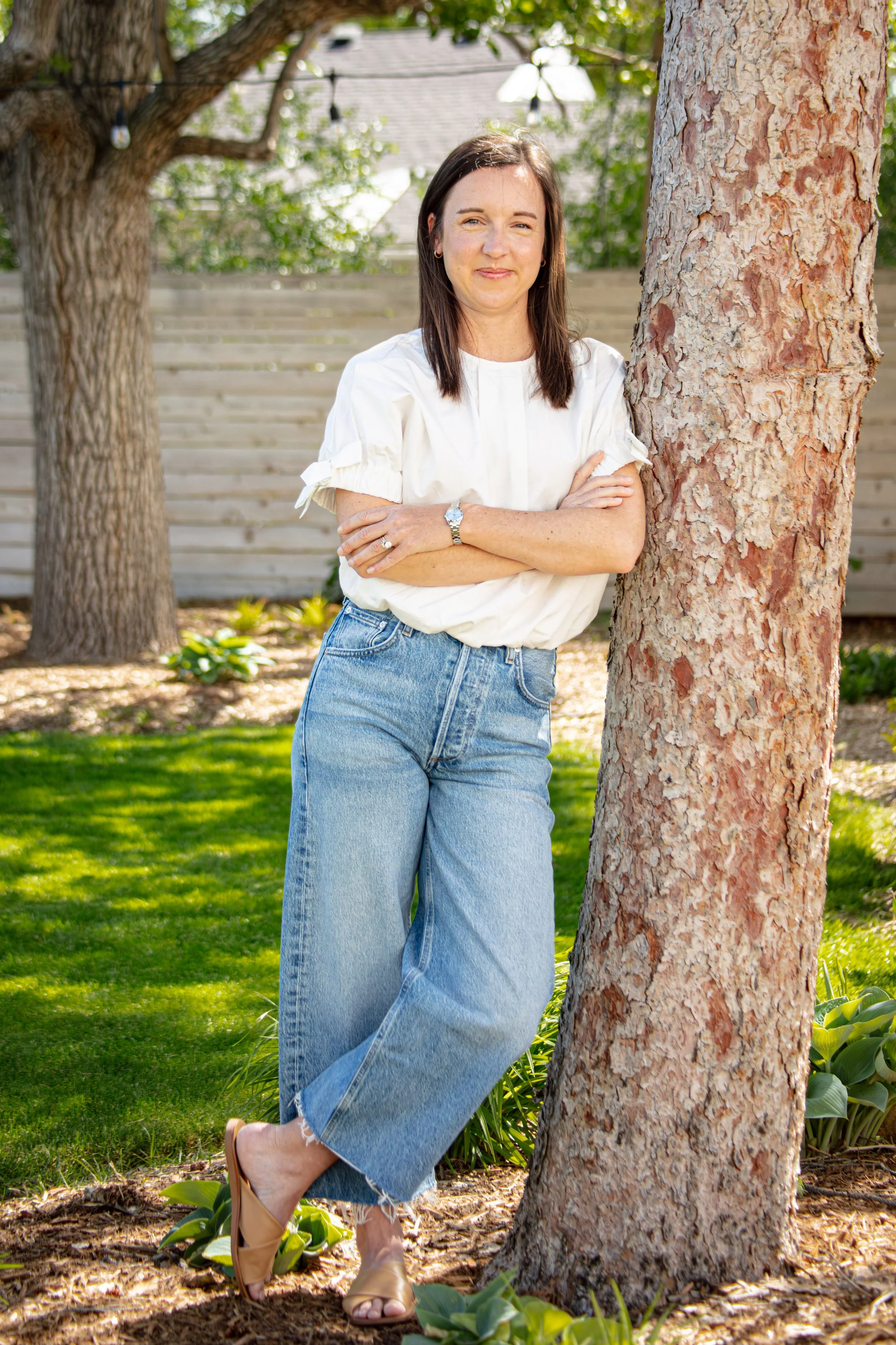 A woman with dark brown hair, wearing a white blouse, blue jeans, and beige high heel sandals, stands outdoors next to a large tree with textured bark. She has crossed arms and is smiling slightly, with a wooden fence, green grass, and leafy plants in the background, and string lights hanging from the tree.