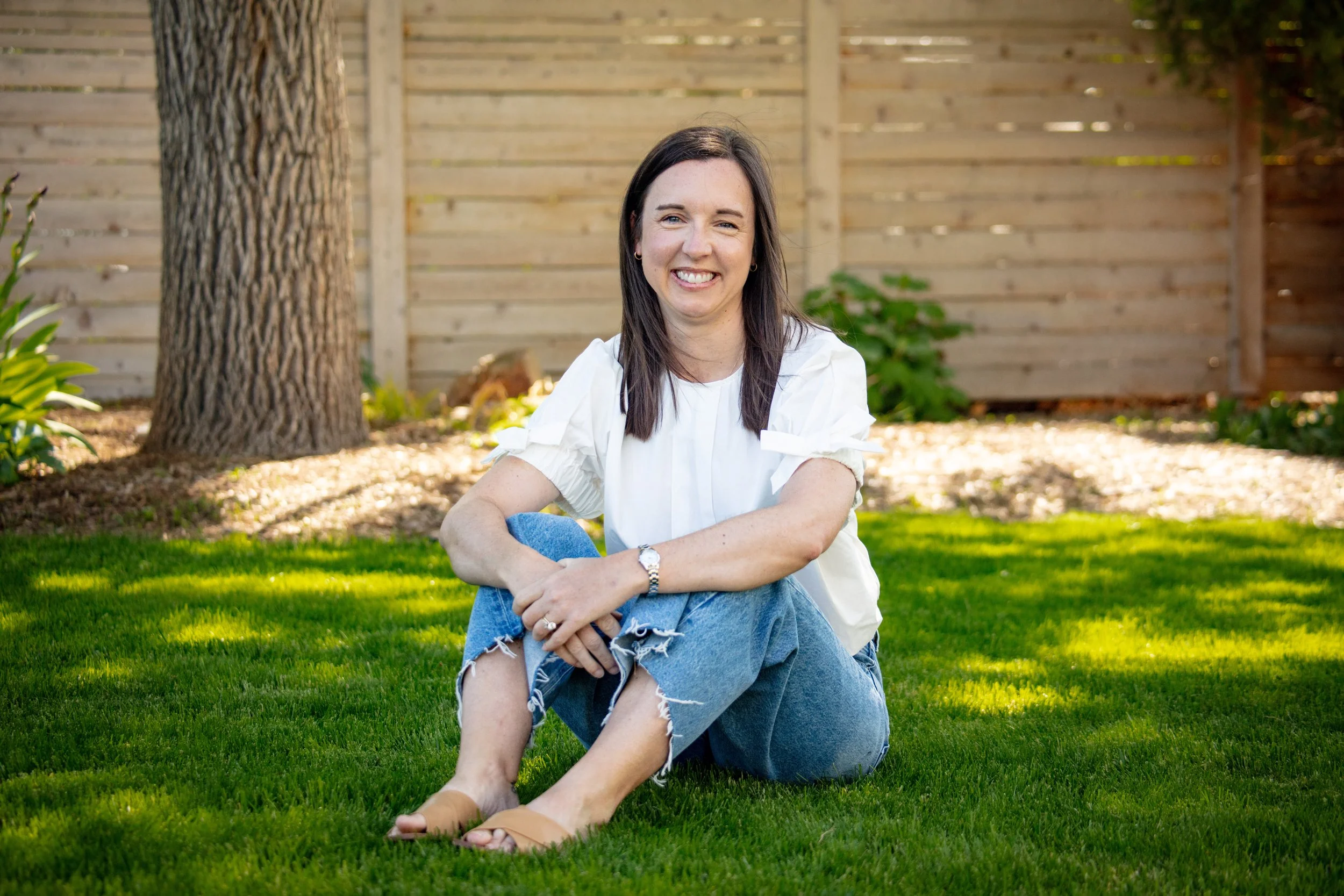 A woman sitting on the grass in a garden, smiling at the camera, with a wooden fence, tree trunk, and green plants in the background.