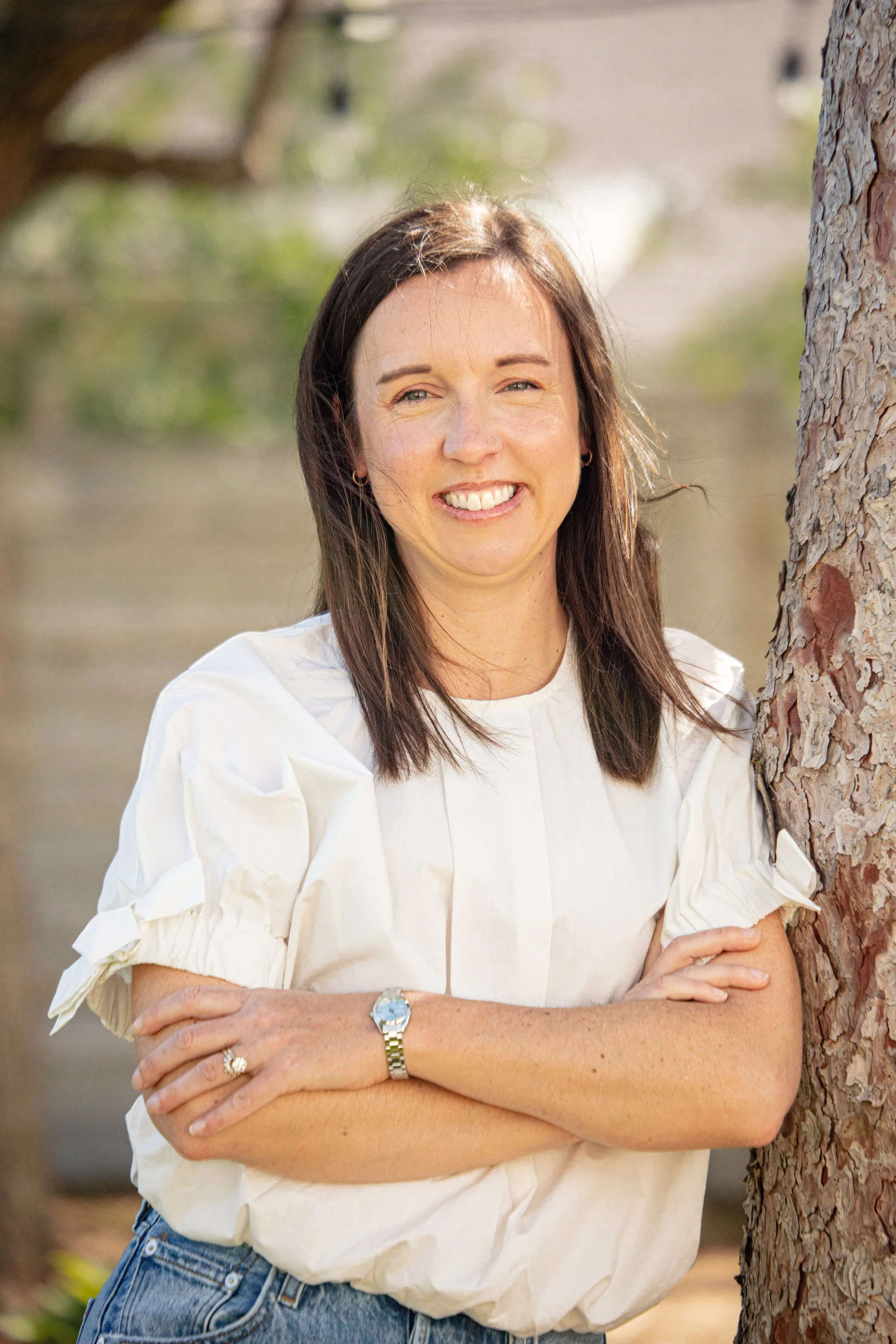A woman standing outdoors next to a tree, smiling and crossing her arms.