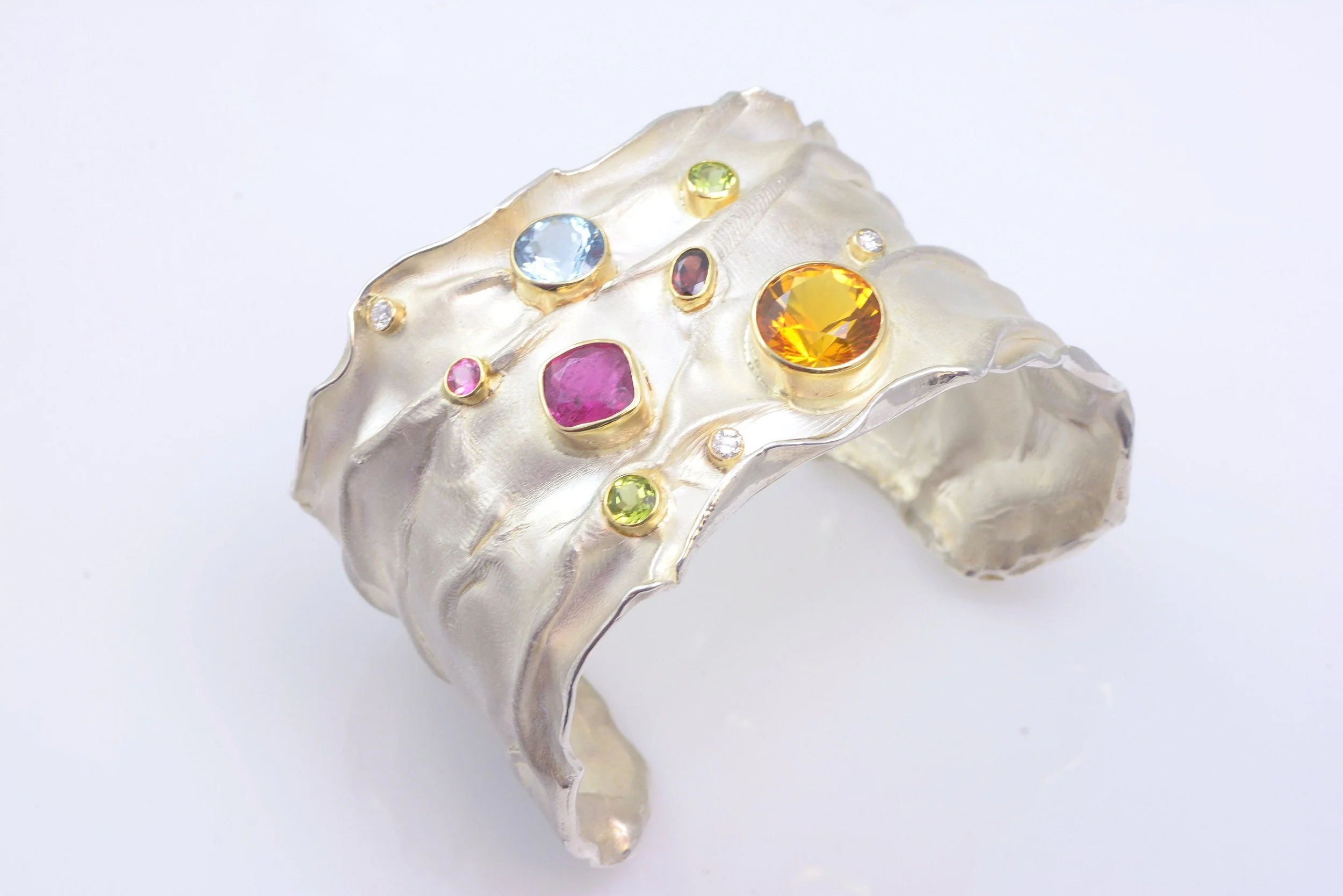 Silver cuff bracelet with multicolored gemstones including pink, blue, yellow, and green stones set in gold. Design by Bill Wismar.