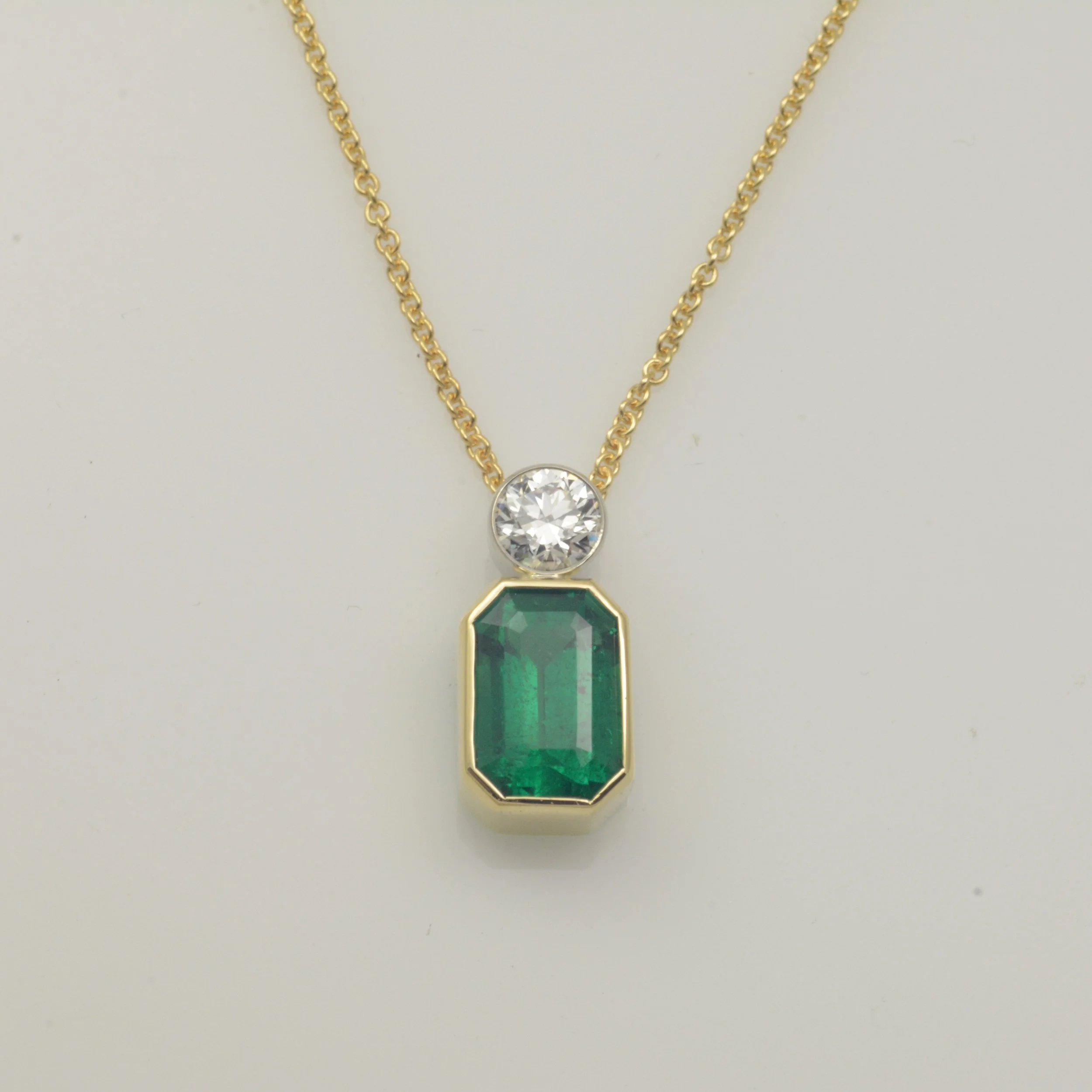 Gold chain necklace featuring a large rectangular emerald gemstone and a smaller round diamond gemstone above it against a white background.