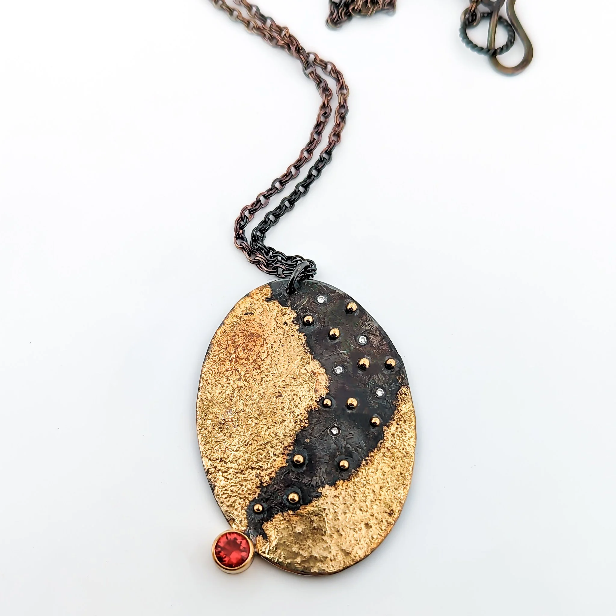 A gold and oxidized black sterling silver oval pendant necklace with a red fire opal gemstone at the bottom, suspended on a chain. Design by Rocky Pardo Jewelry.