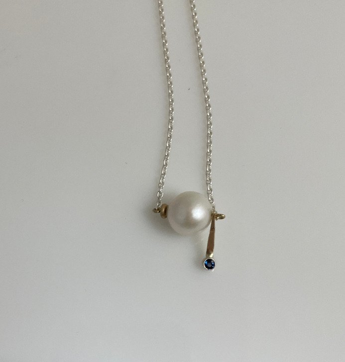 Silver necklace with a large pearl and a vertical bar pendant with a small blue sapphire gemstone. Design by Bill Wismar.