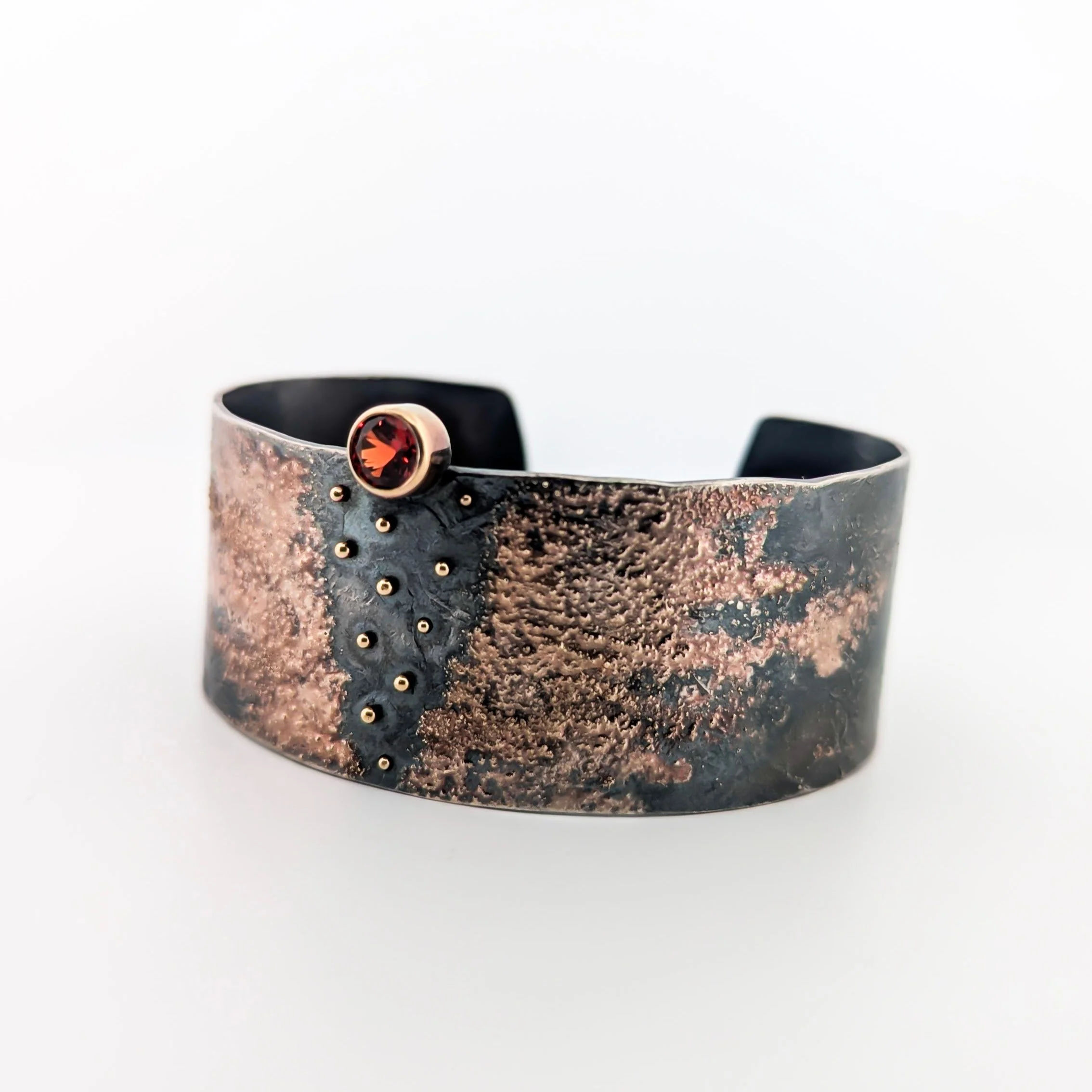 An oxidized sterling silver bracelet with a black and gold textured design, decorated with red garnet gemstone at the top and tiny kinetic gold dots. Design by Rocky Pardo.