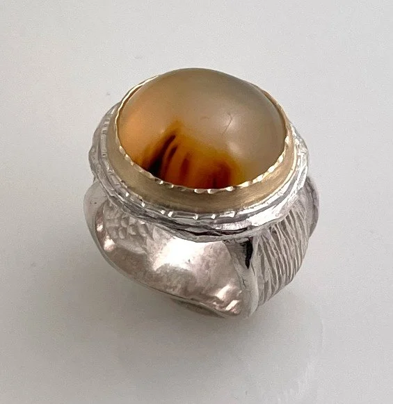 A silver ring with a large beige and brown agate stone set in it, viewed from above on a plain white background. Design by Crazy Hair Susan.