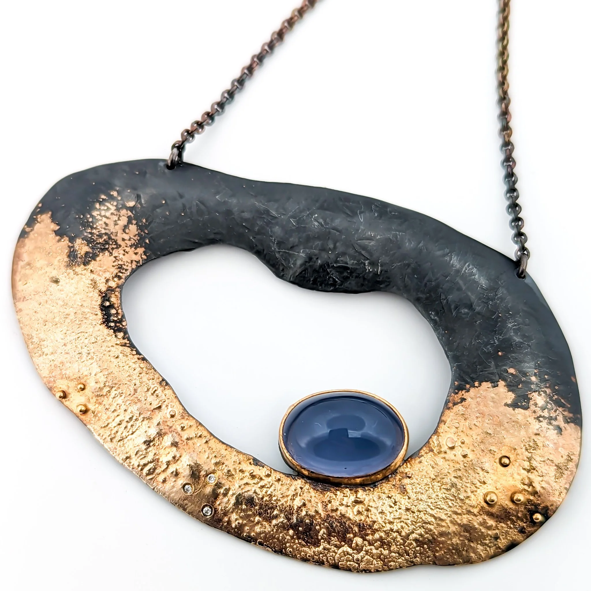 A large statement necklace with an irregular oval shape, featuring oxidized black sterling silver and gold textured sections, and a round blue chalcedony gemstone in a gold bezel at the bottom center, hanging from a dark chain. Design by Rocky Pardo.