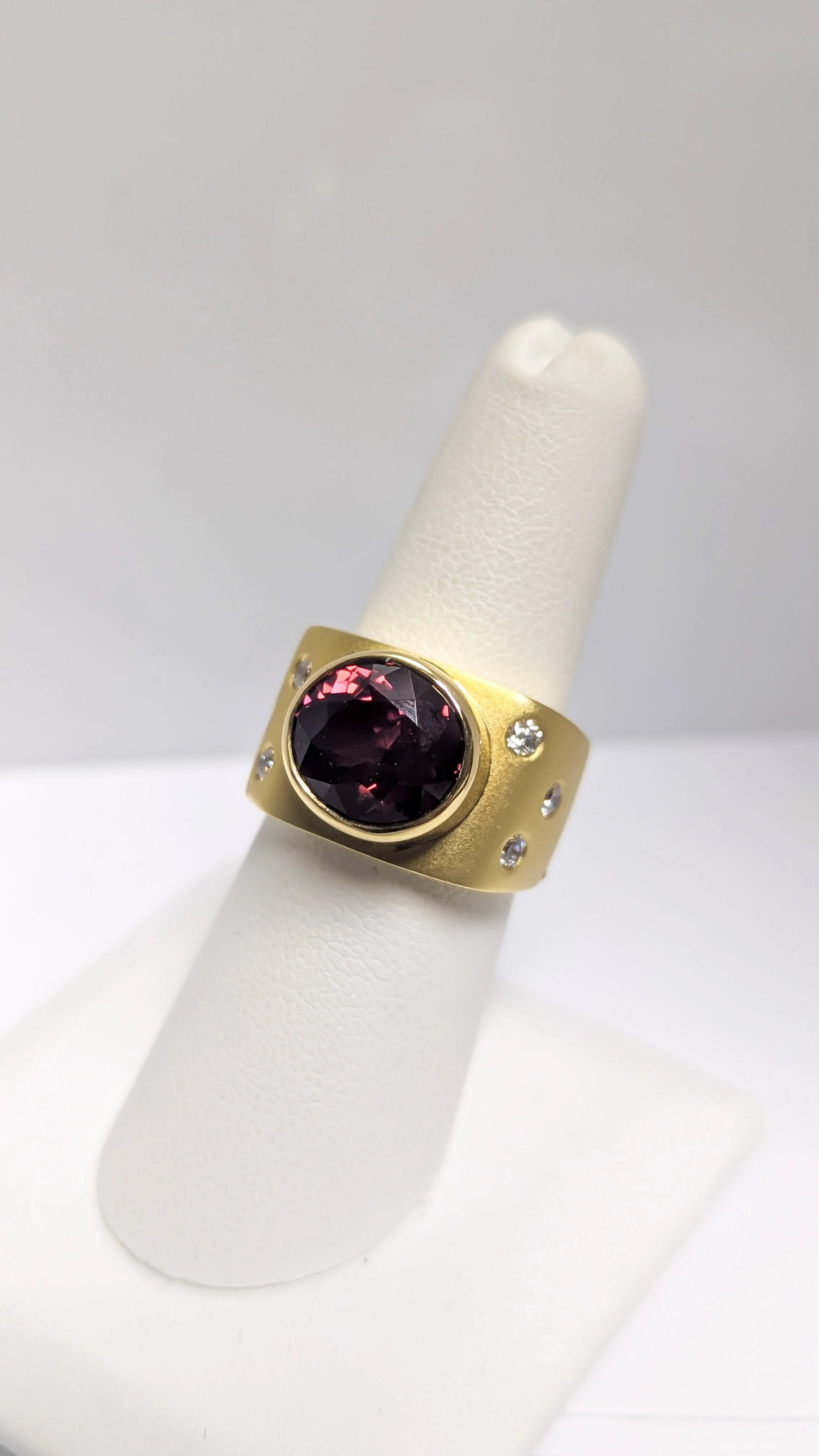 Gold ring with large round dark red Rhodiolite garnet gemstone and diamonds, displayed on a white ring holder. Design by Bill Wismar.