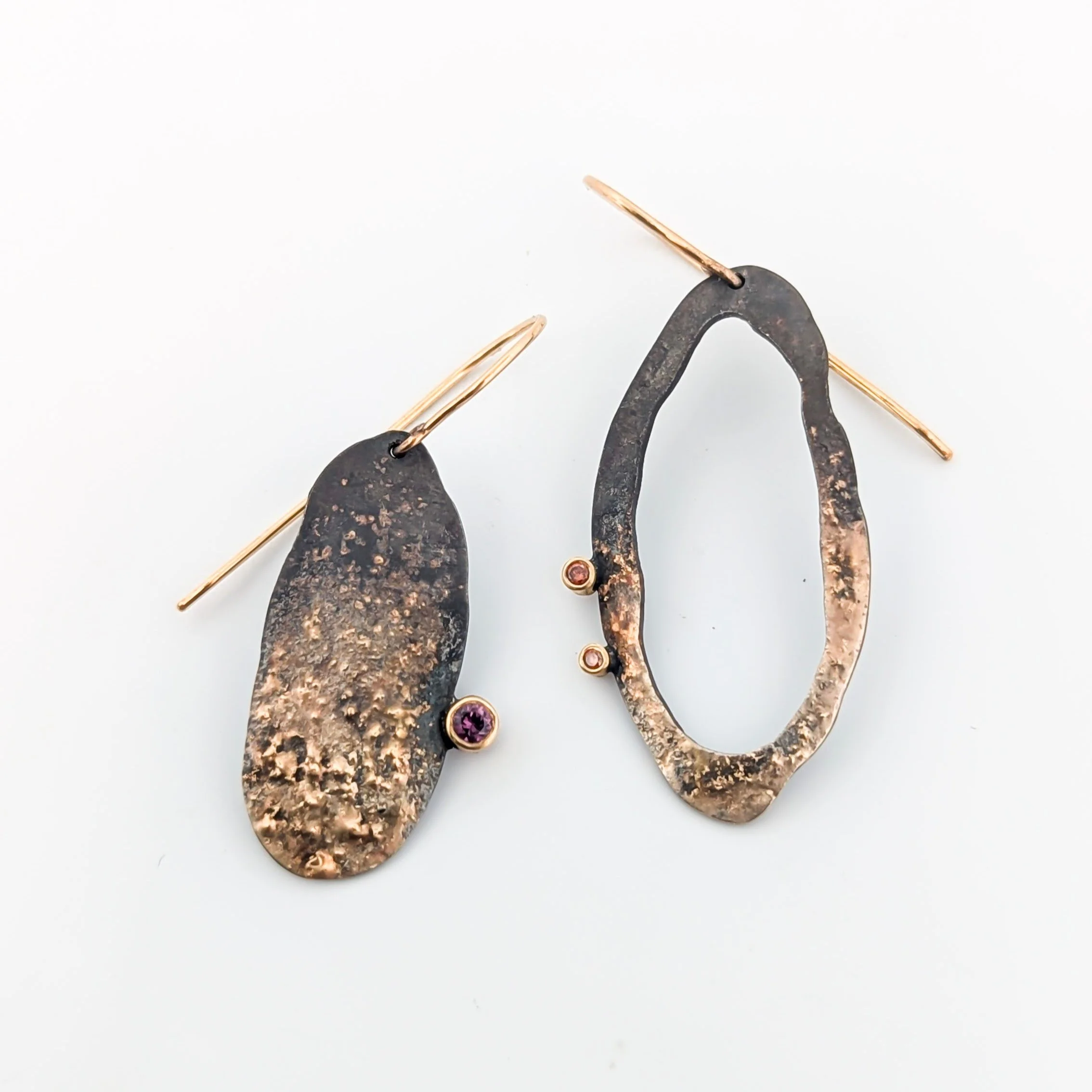 Pair of artistic drop-shaped earrings with hammered metal texture and small sapphire and diamond gemstones, one with a purple stone and the other with a pinkish stone. Oxidized sterling silver and gold. Design by Rocky Pardo Jewelry.