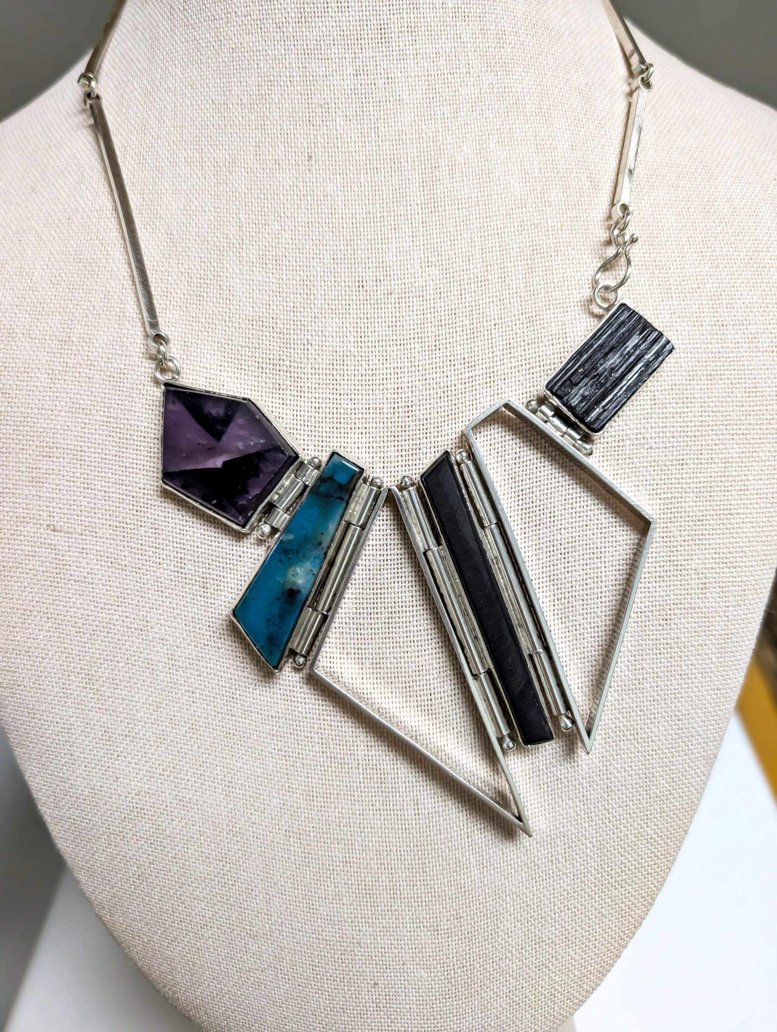 A statement necklace with geometric shapes and colorful stones displayed on a beige bust form. Created by Dirigible Designs.