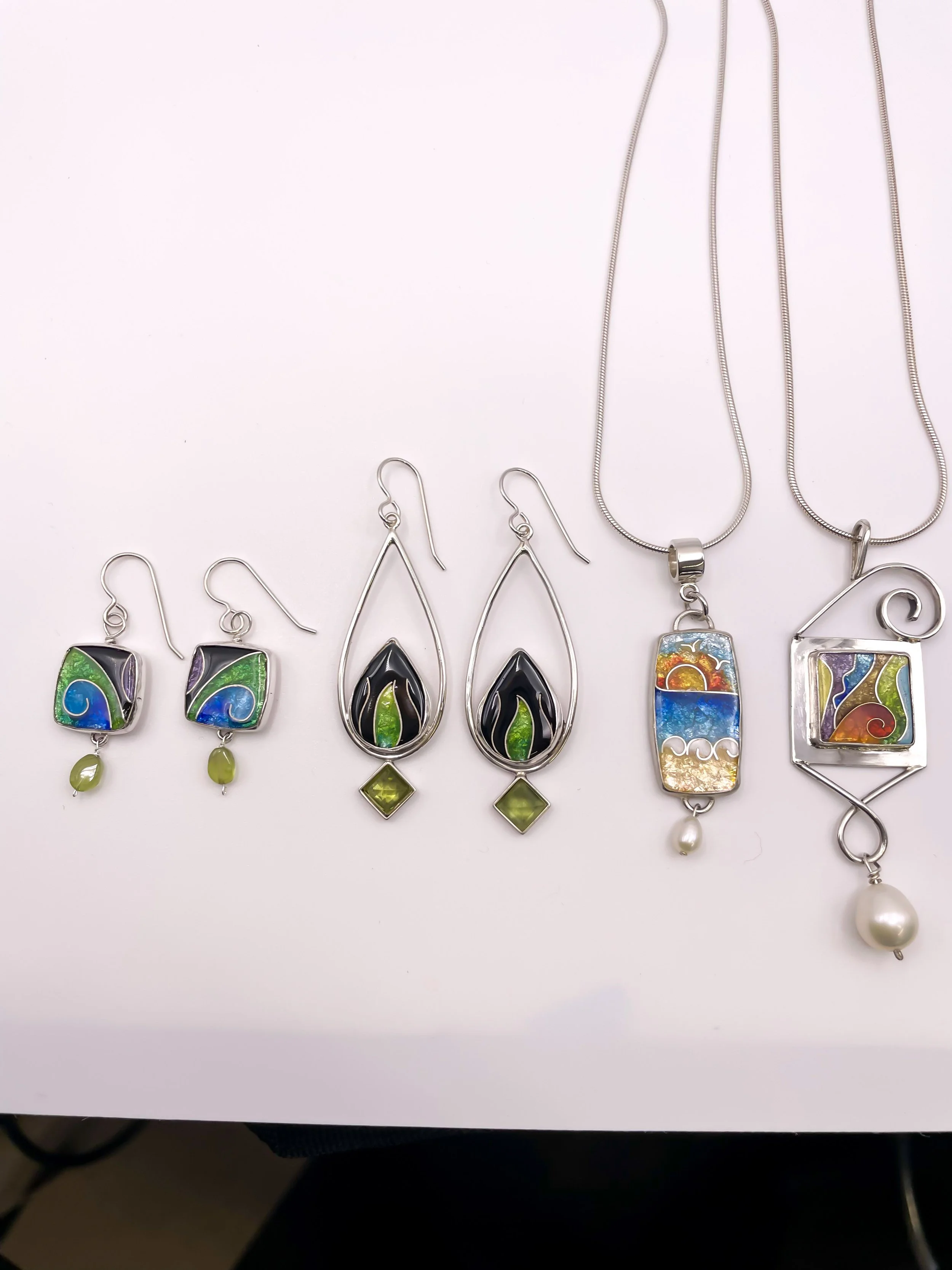 Set of five colorful jewelry pieces, including earrings and necklaces, with abstract and floral designs in silver and vibrant colors, some with beads or pearls using cloissonne enamel technique. Designs by Vitrice McMurry.