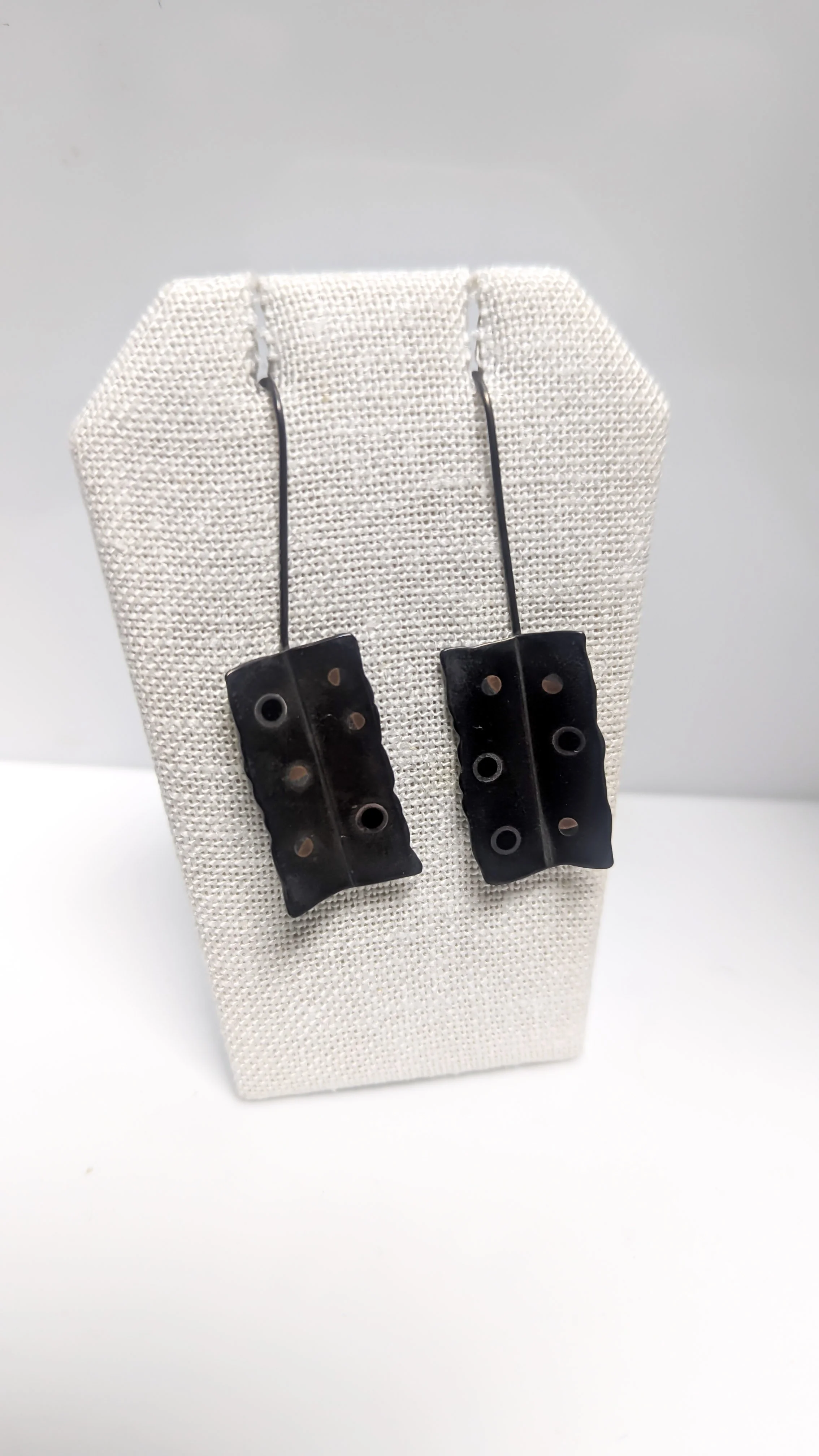 A pair of black earrings with metal hooks displayed on a beige fabric jewelry stand. Uses Japanese alloys and shakudo techniques. Design by J. Collier.