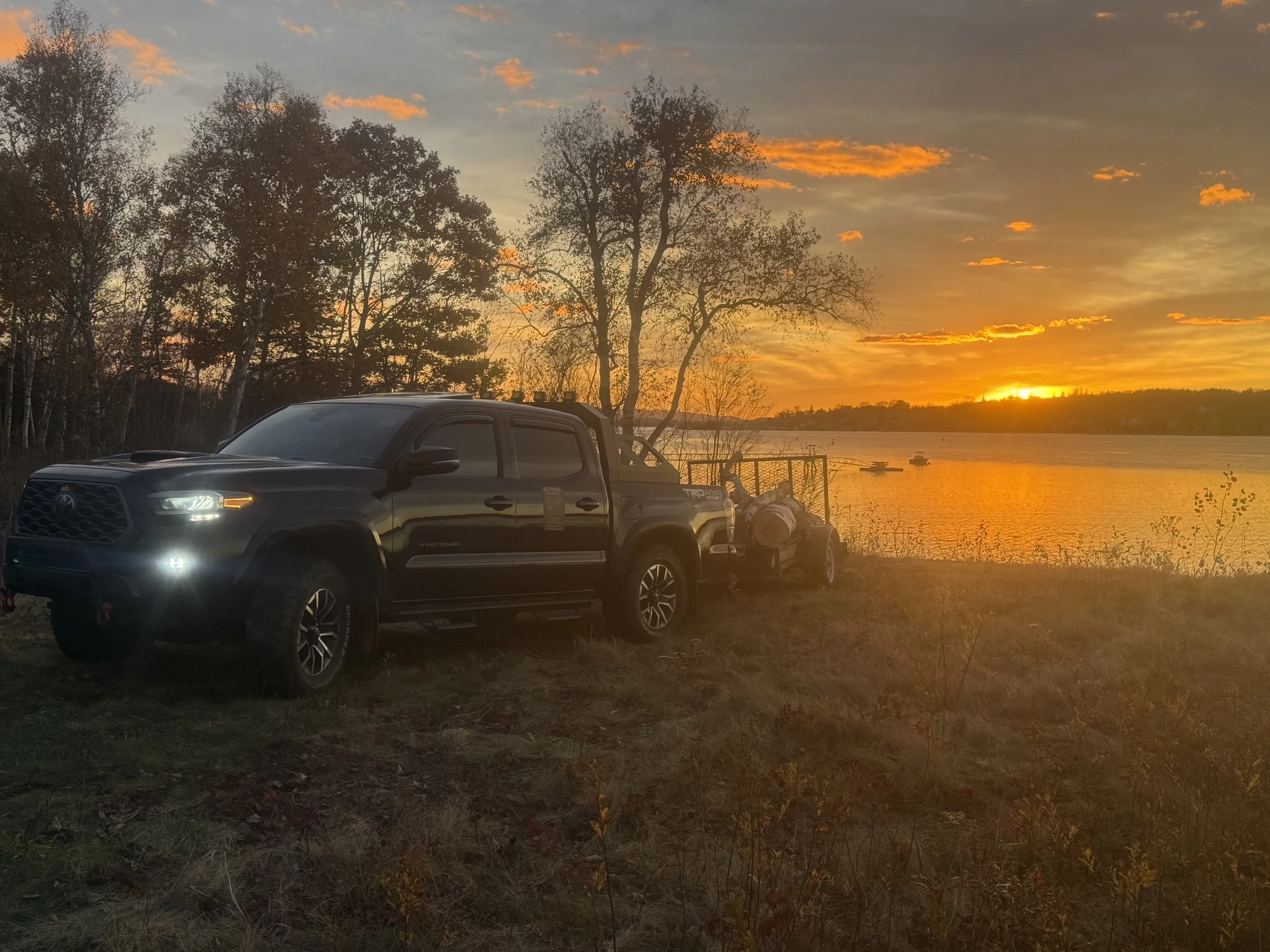 A black Toyota pickup truck parked near a lake at sunset with trees in the background and boats on the water.