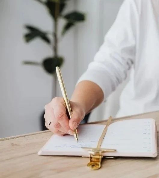 A person wearing a white shirt writing in a planner with a gold pen, on a wooden desk with a green plant in the background.