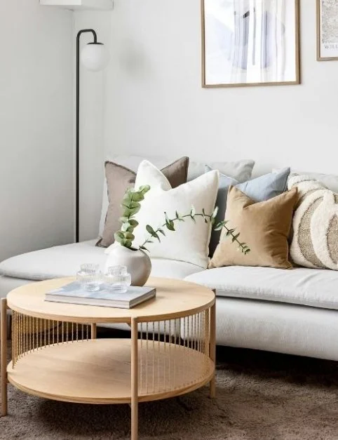 Living room with a white sofa, beige and blue pillows, and a round wooden coffee table with a vase holding green eucalyptus, glassware, and books. Floor lamp and framed artwork on the wall.