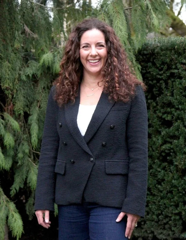 A woman with curly brown hair smiling outdoors in front of greenery, wearing a dark blazer and white top.