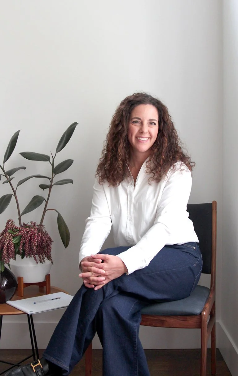 A woman with curly brown hair smiling and sitting on a wooden chair next to a small table with a notebook and pen. There is a potted plant with large green leaves and pinkish-red flowers on the table. The background is a plain white wall.