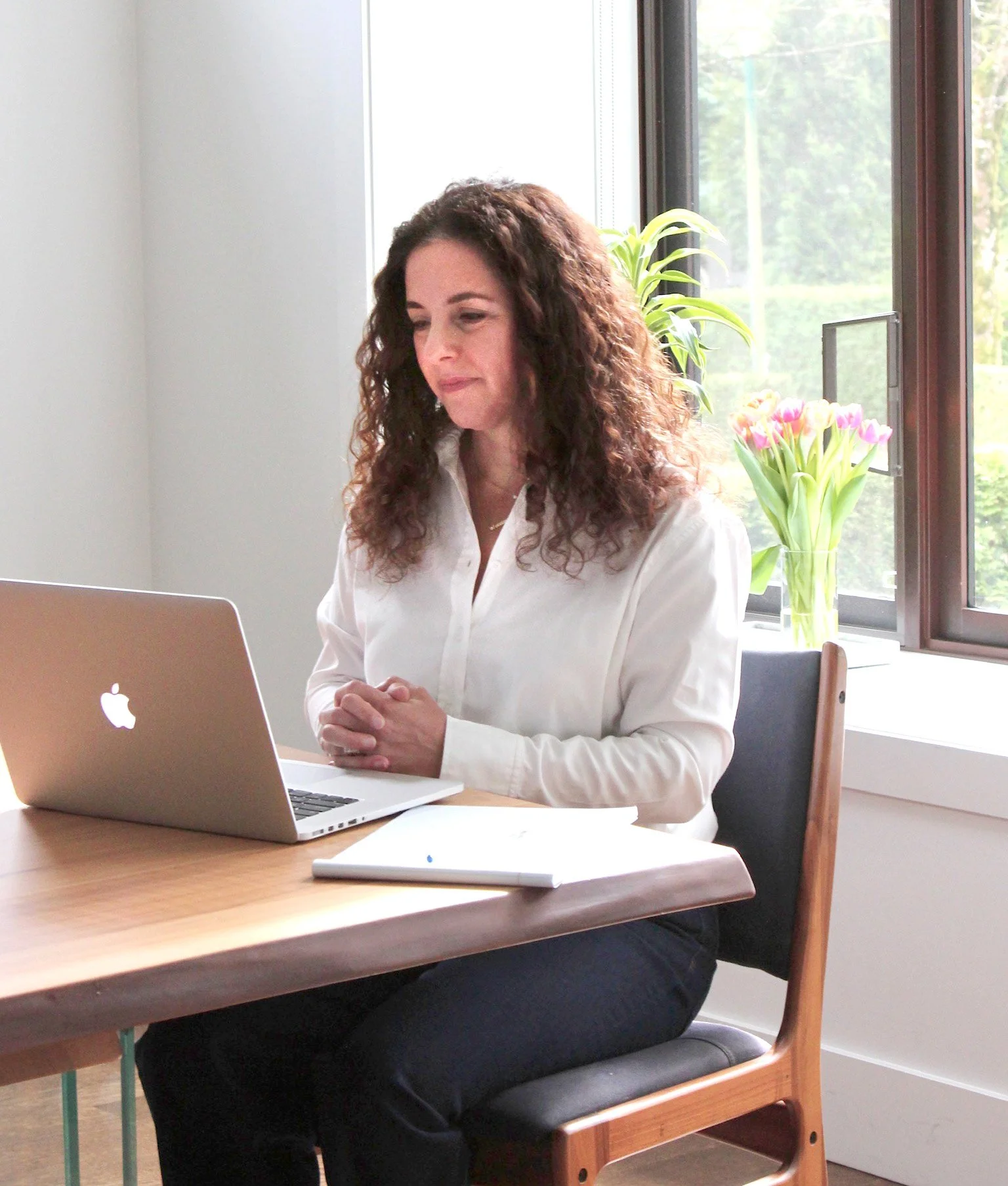 A woman with curly brown hair sits at a wooden table, working on a silver MacBook laptop. She is wearing a white blouse and dark pants. In the background, there is a window with a view of trees, and a glass vase of pink and yellow tulips on the windowsill.