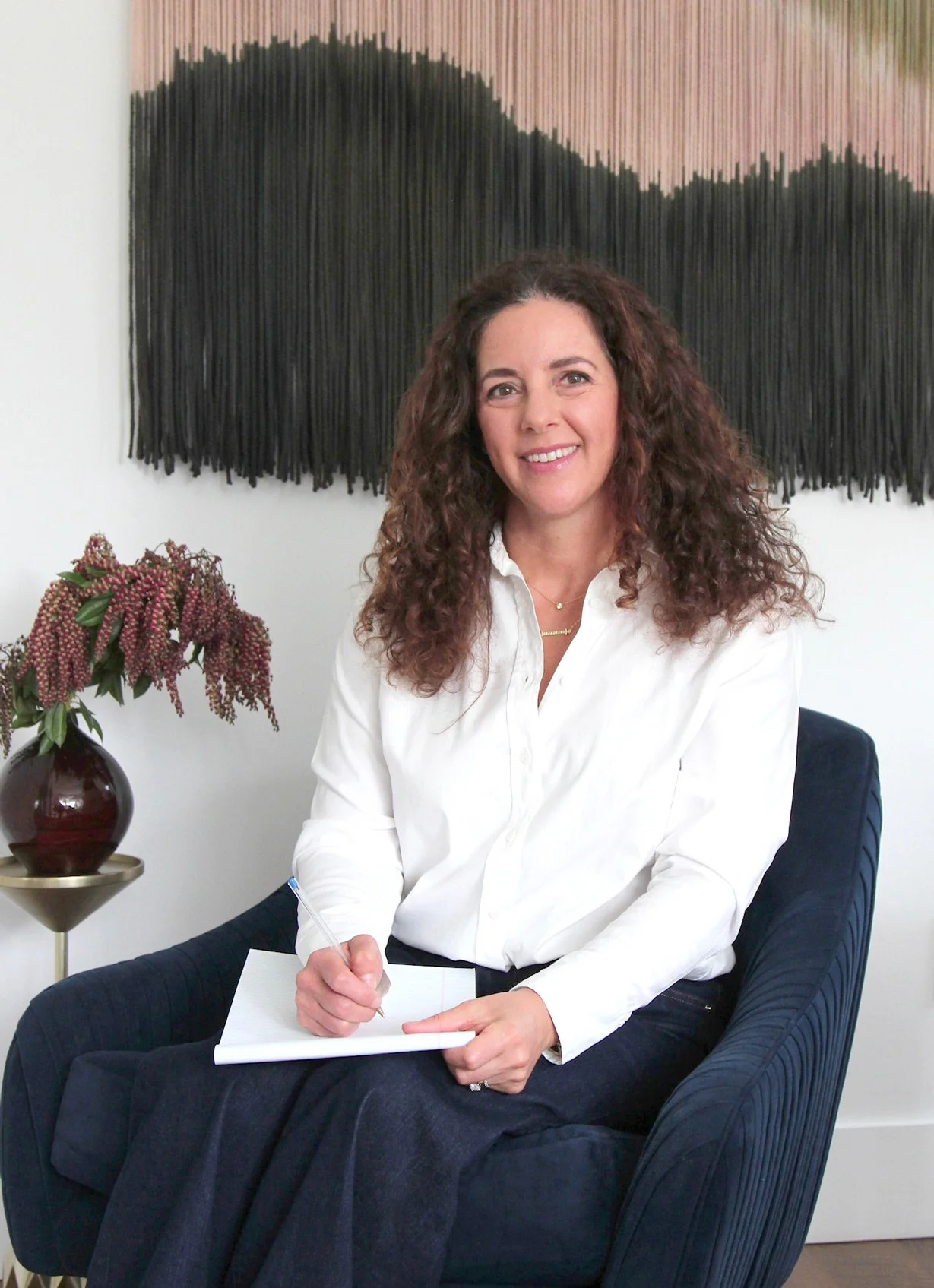A woman with curly brown hair and a white blouse sitting in a modern navy armchair, holding a pen and writing on a notepad. Behind her is a large black textured artwork on the white wall, and to her left is a small round table with a dark vase holding pinkish flowers.