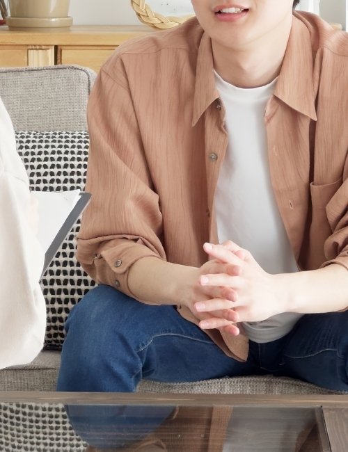 Person sitting on a sofa with hands clasped, wearing a tan shirt over a white t-shirt, in a cozy, well-lit room.