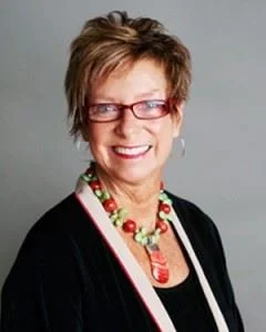 A woman with short brown hair, wearing glasses, a black top, and colorful jewelry, smiling at the camera.