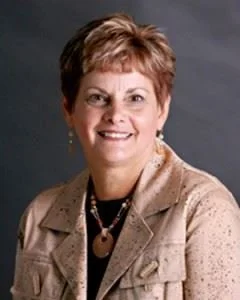 A woman with short reddish-brown hair smiling, wearing a beige jacket and a necklace, against a dark background.
