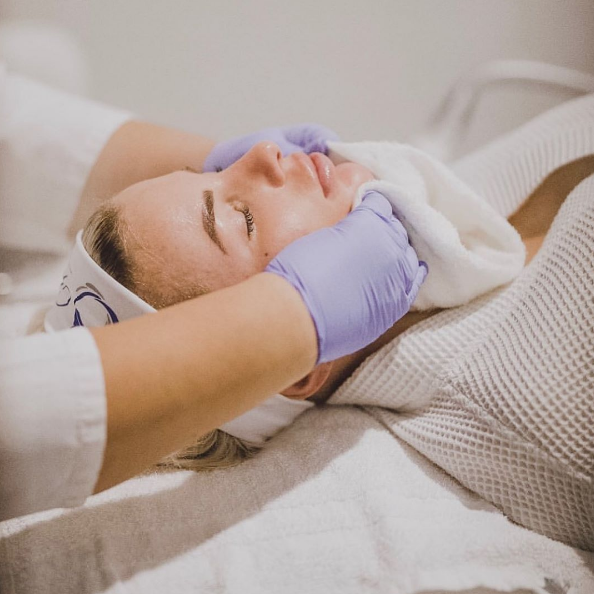 A woman receiving a facial treatment from a skincare professional wearing purple gloves, lying down with her eyes closed.