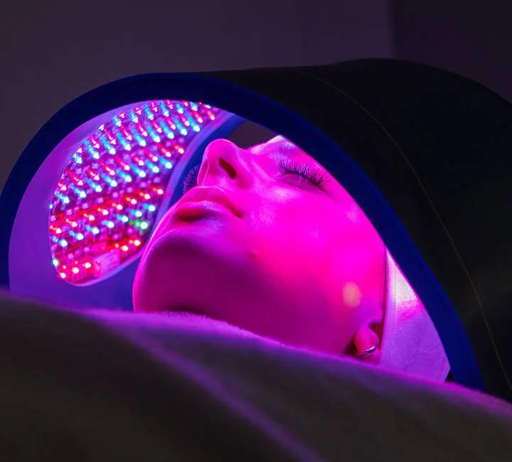 A person receiving a facial treatment under an LED light mask emitting vibrant pink and purple lights, with the person's eyes closed during the procedure.
