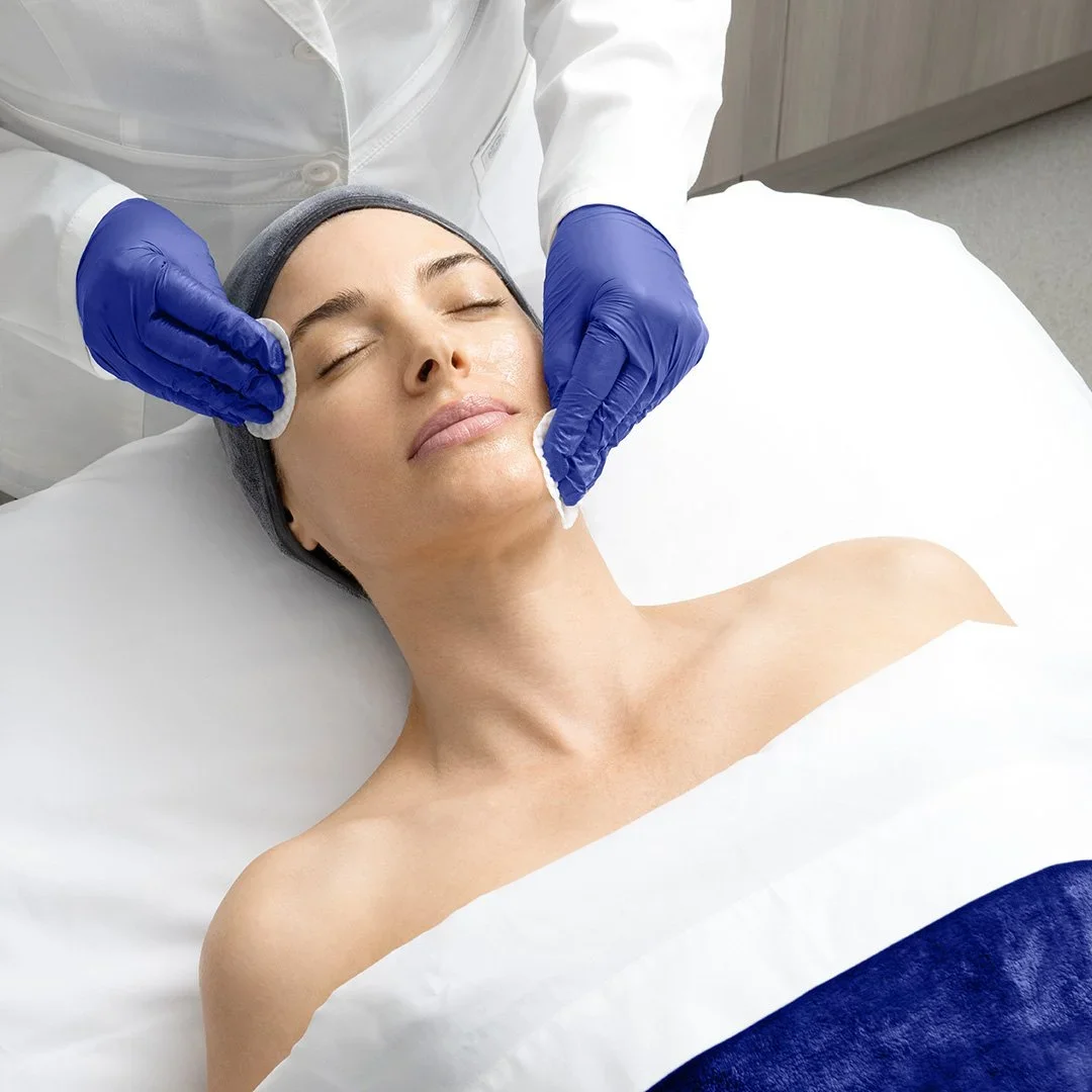 A woman receiving a facial treatment from a professional wearing blue gloves at a spa or clinic setting.
