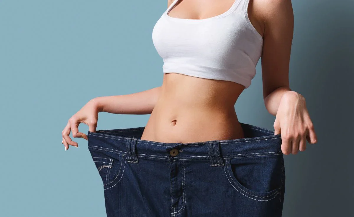 A woman wearing a white crop top and loose jeans, holding the waistband away to show she has lost weight.