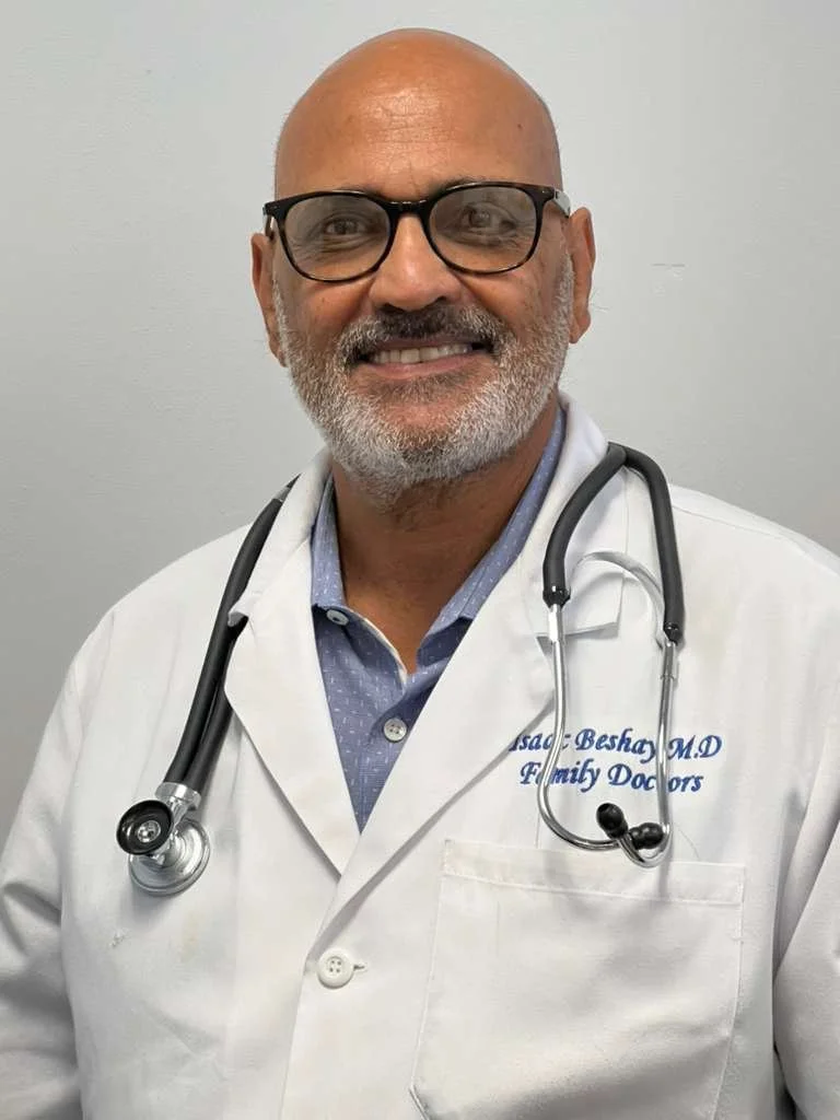 A smiling male doctor with glasses, a gray beard, and a stethoscope around his neck, wearing a white lab coat with embroidered text, standing against a plain light gray wall.