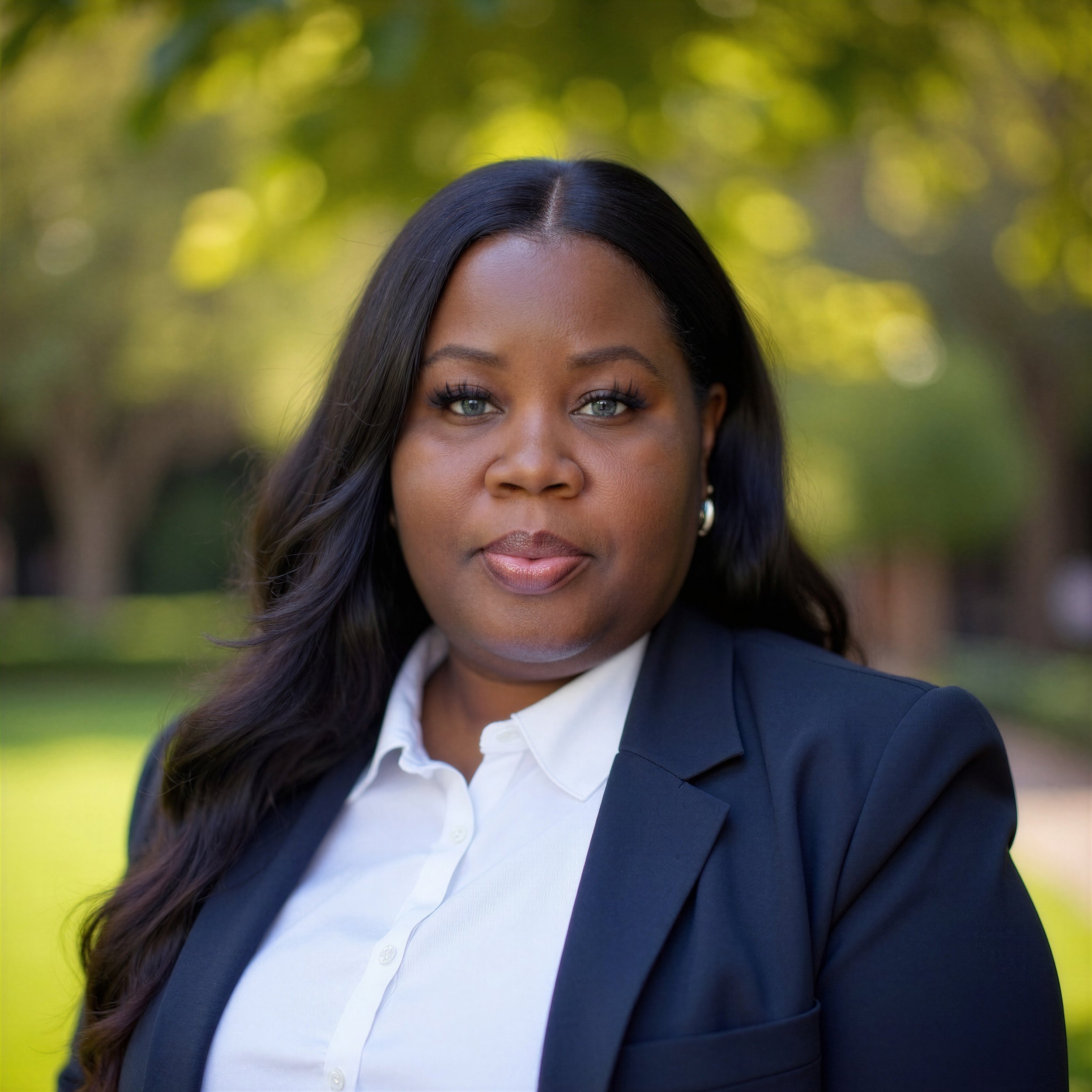 A portrait of an African American woman with long dark hair, dressed in a white blouse and navy blazer, standing outdoors with trees and greenery in the background.