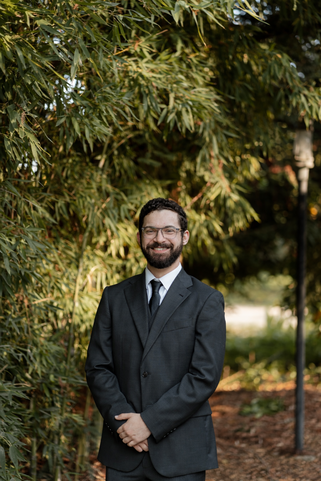 A man in a black suit, white shirt, black tie, and glasses, smiling and standing outdoors in front of green foliage.