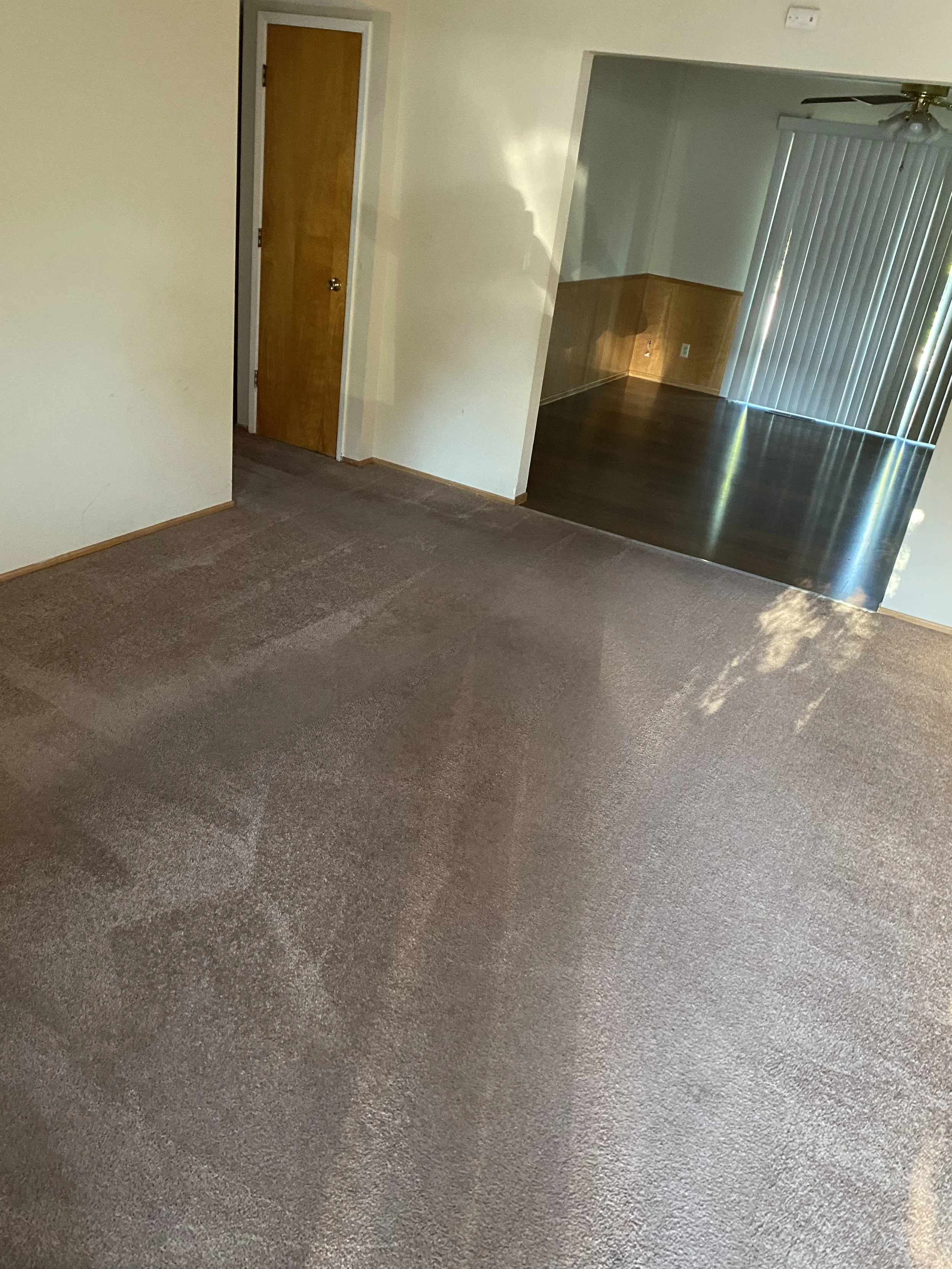 Empty living room with beige carpet, open doorway leading to a room with dark wood flooring, wood paneled wall, sliding glass door with vertical blinds, and a ceiling fan.