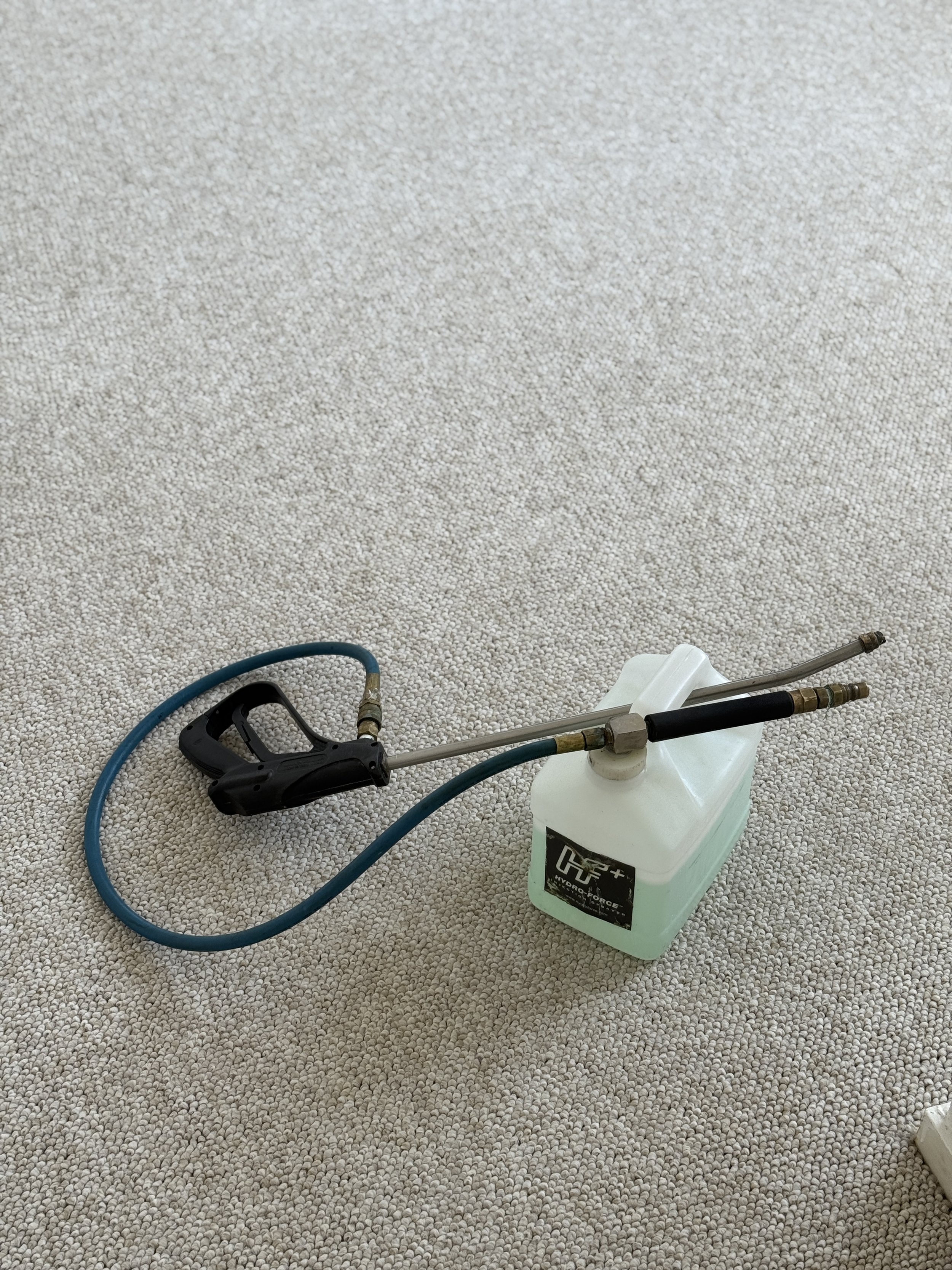 Electric pressure washer on beige carpeted floor with a spray gun and hose connected to a white container.