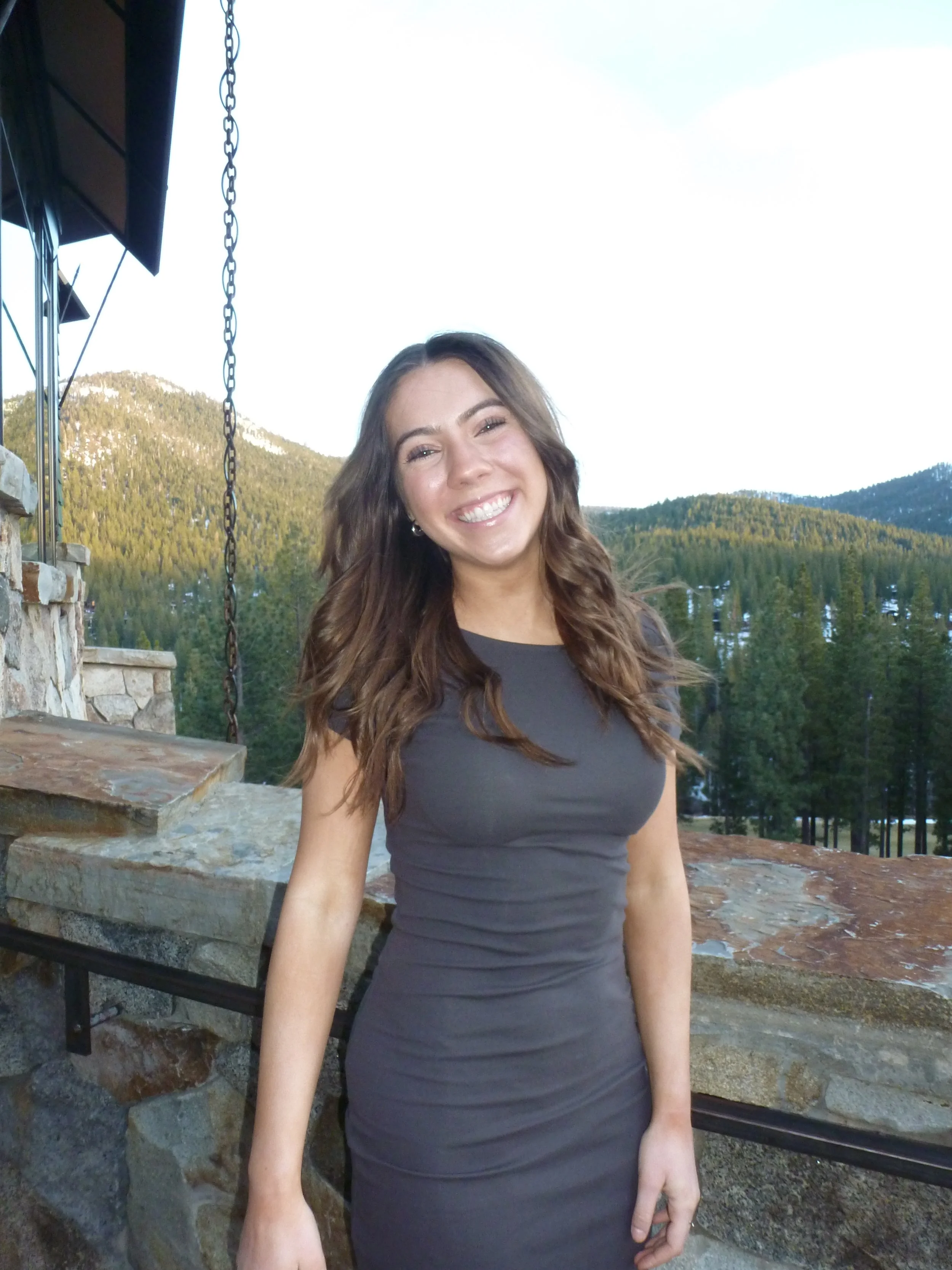 A smiling woman with long brown hair wearing a black dress, standing outdoors with mountains and trees in the background.