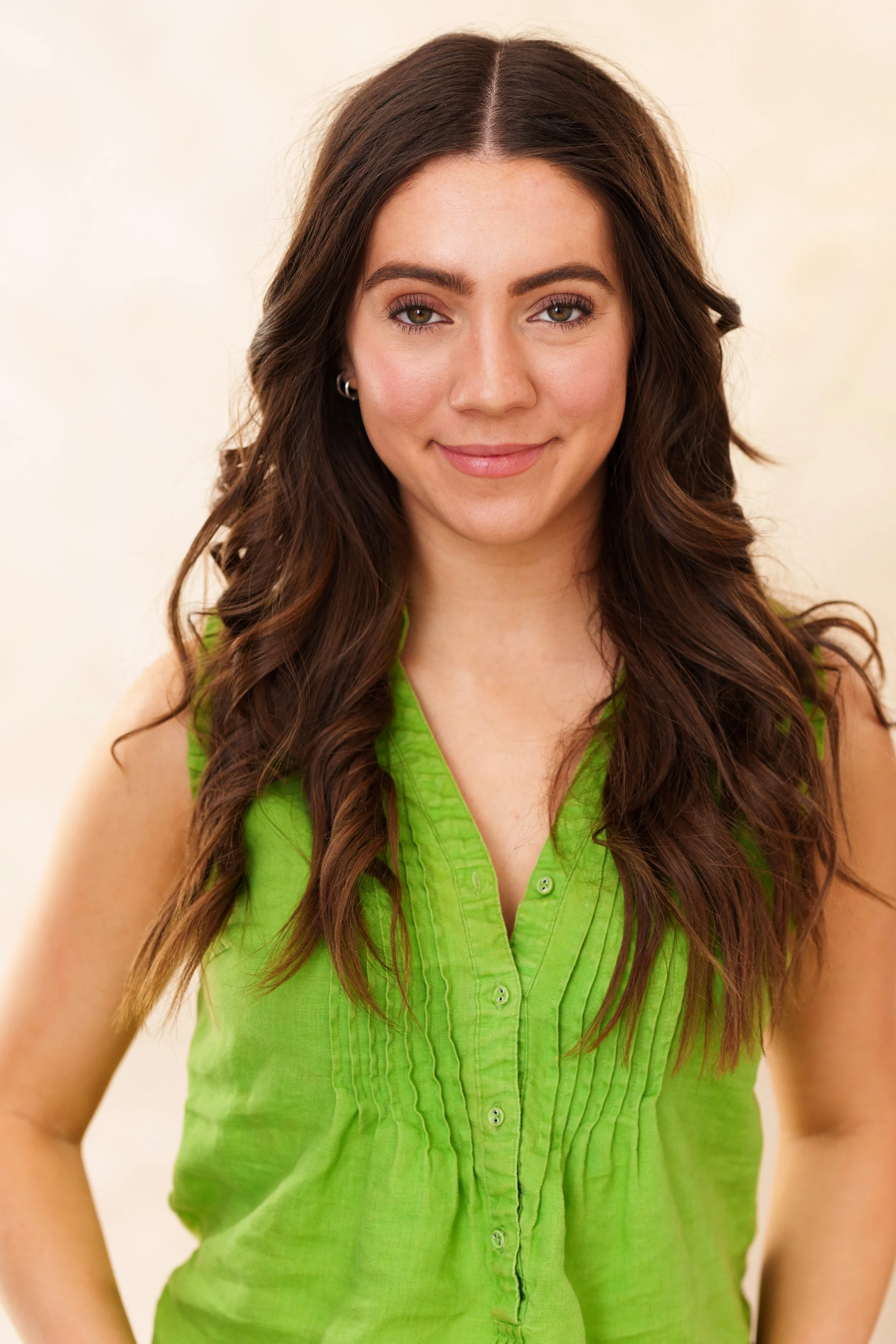 A young woman with long, wavy brown hair and a light complexion, wearing a sleeveless lime green blouse, smiling at the camera.