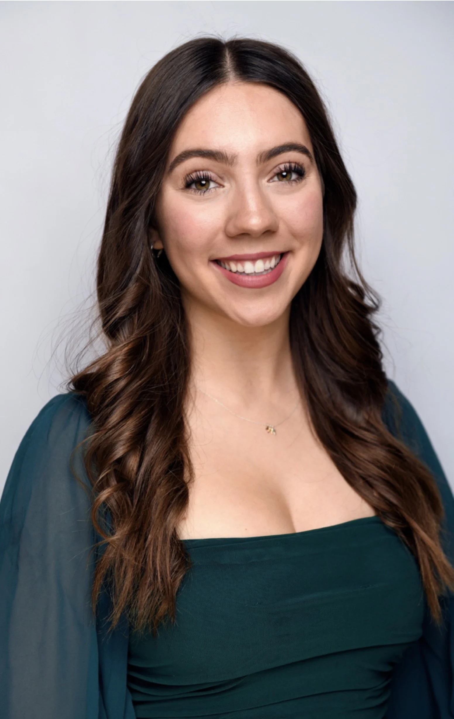 A smiling young woman with long wavy brown hair, wearing a dark green top and a delicate necklace, standing against a plain light gray background.