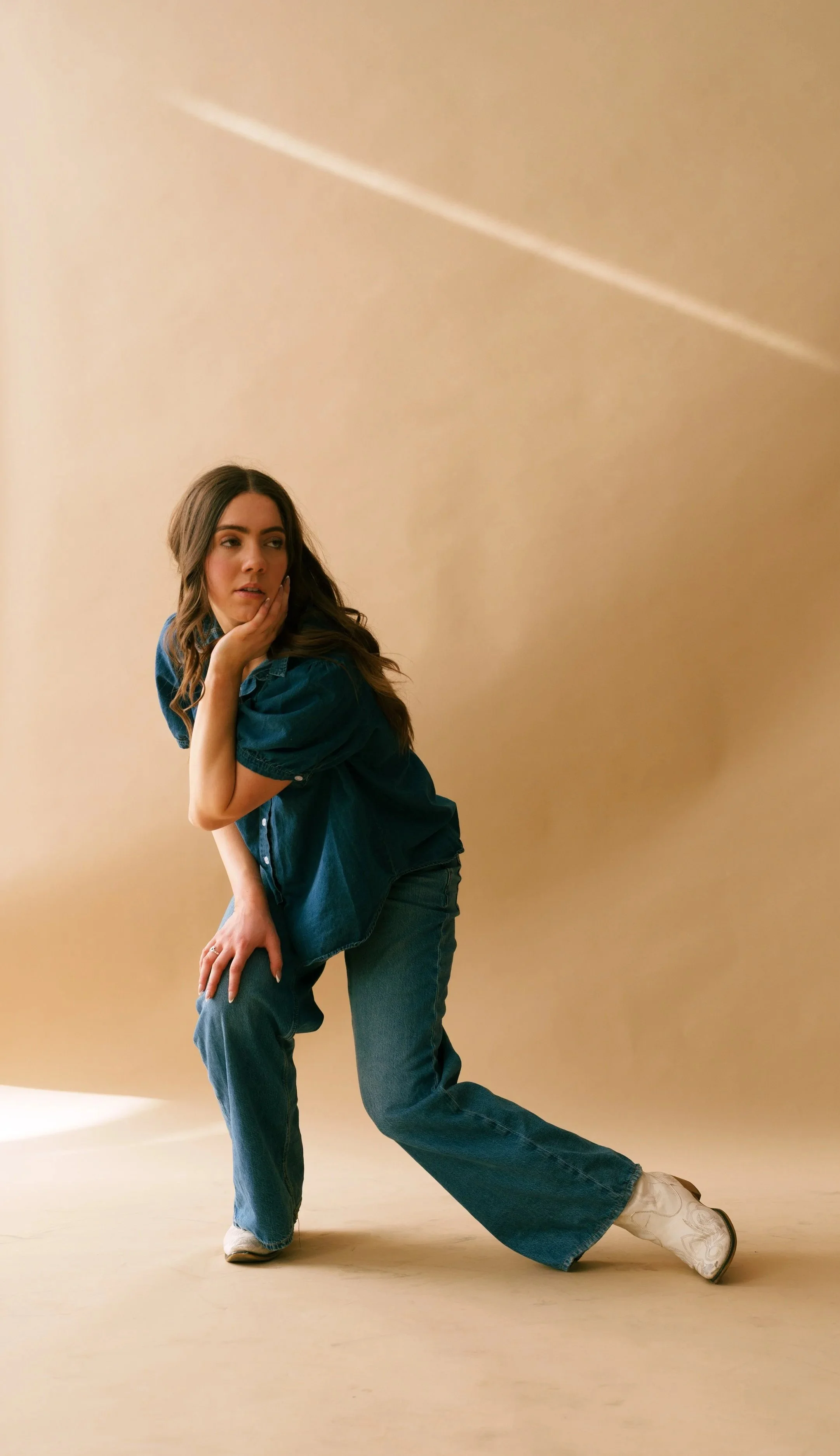 A woman with long brown hair posing against a plain beige background, wearing a loose denim shirt, baggy jeans, and white cowboy boots, with her chin resting on her hand.