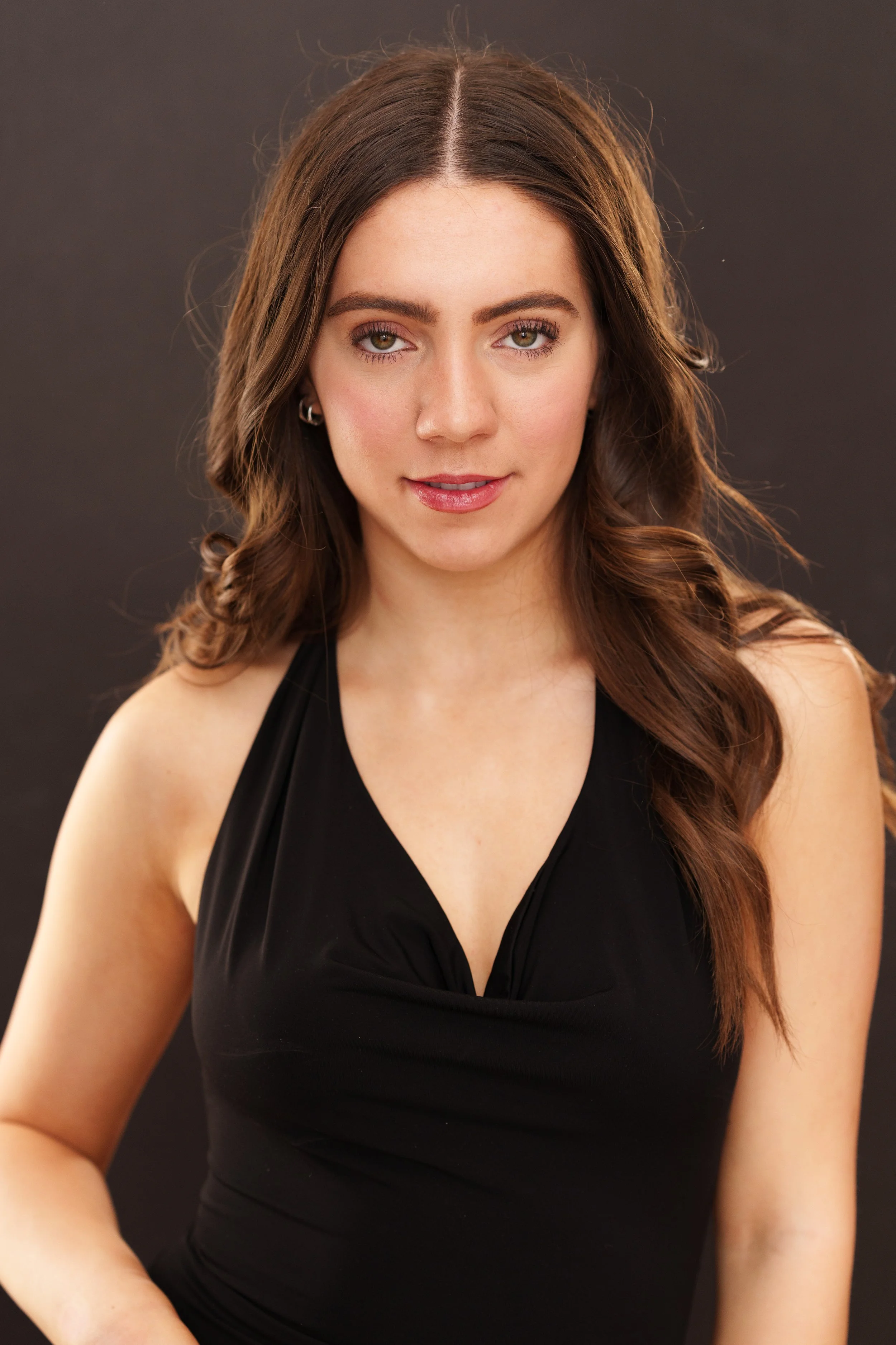 A young woman with long wavy brown hair, wearing a black sleeveless top, is looking directly at the camera with a neutral expression in a studio setting with a dark background.