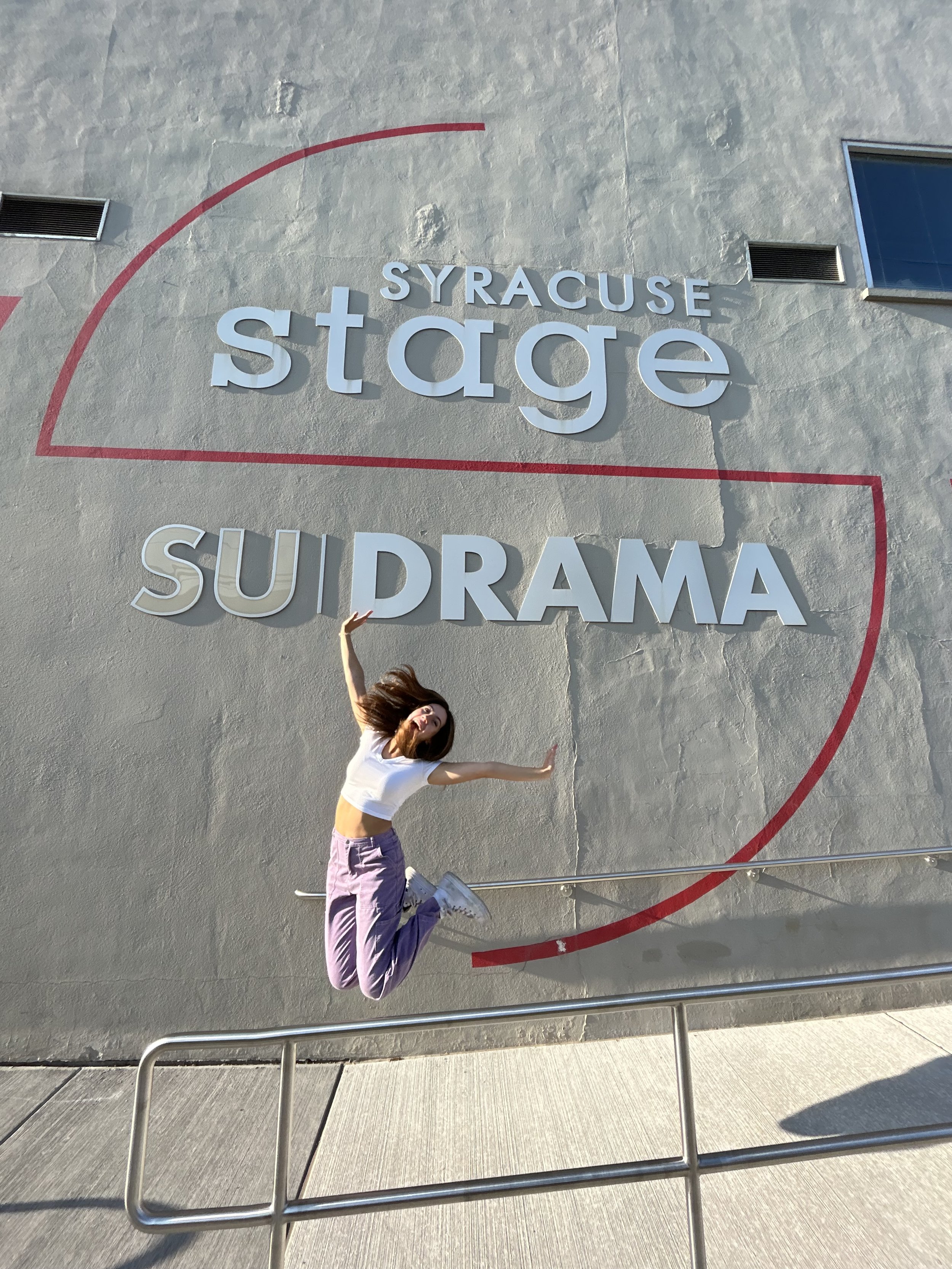 A woman jumping in front of a large wall sign that reads 'SYRACUSE stage SUIDRAMA'. The wall is gray with some texture, and the woman appears to be happy, mid-jump with arms raised and hair in motion, wearing a white crop top, purple pants, and white sneakers.